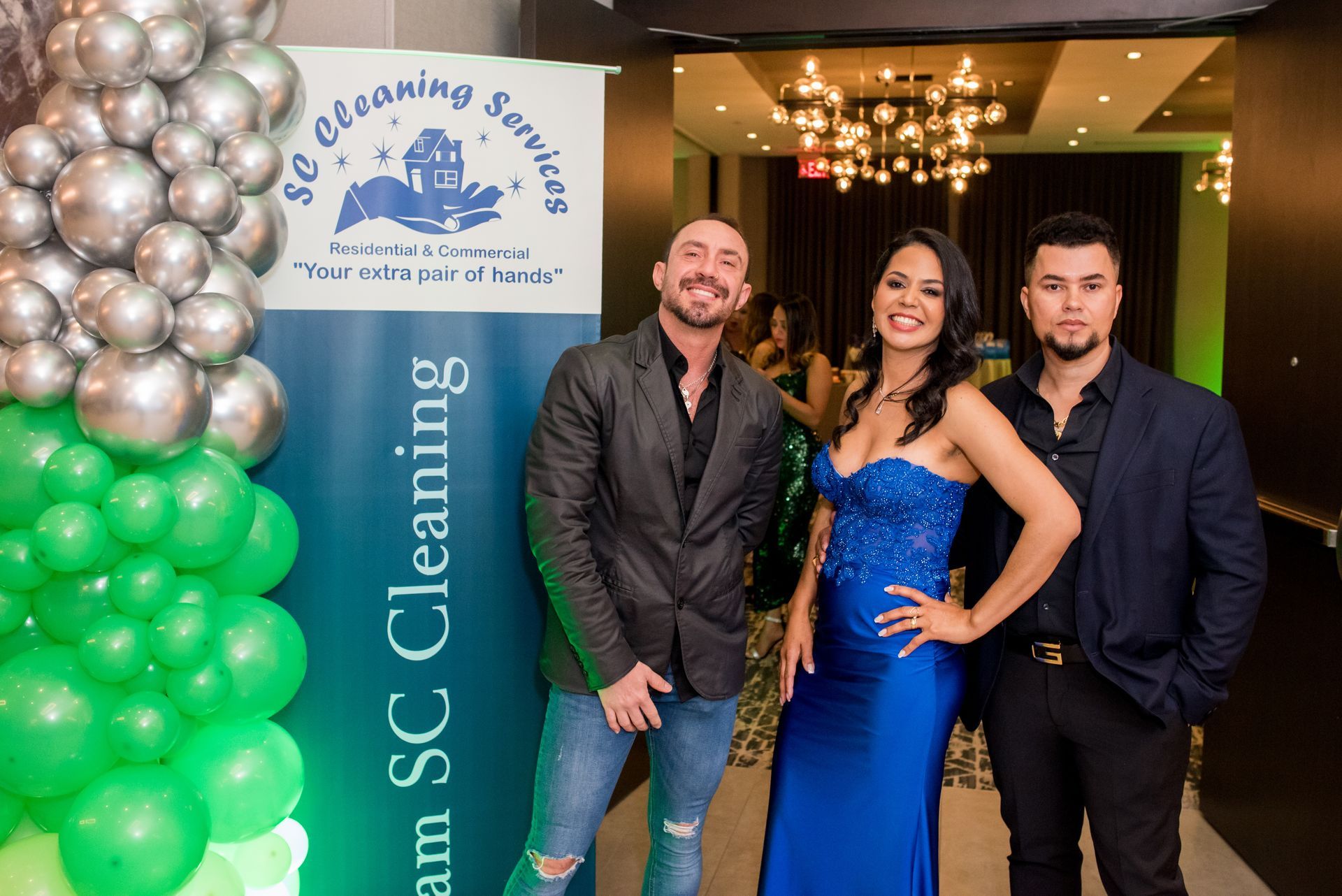 Three people pose in front of a banner. Woman in blue dress, two men, green and silver balloons.
