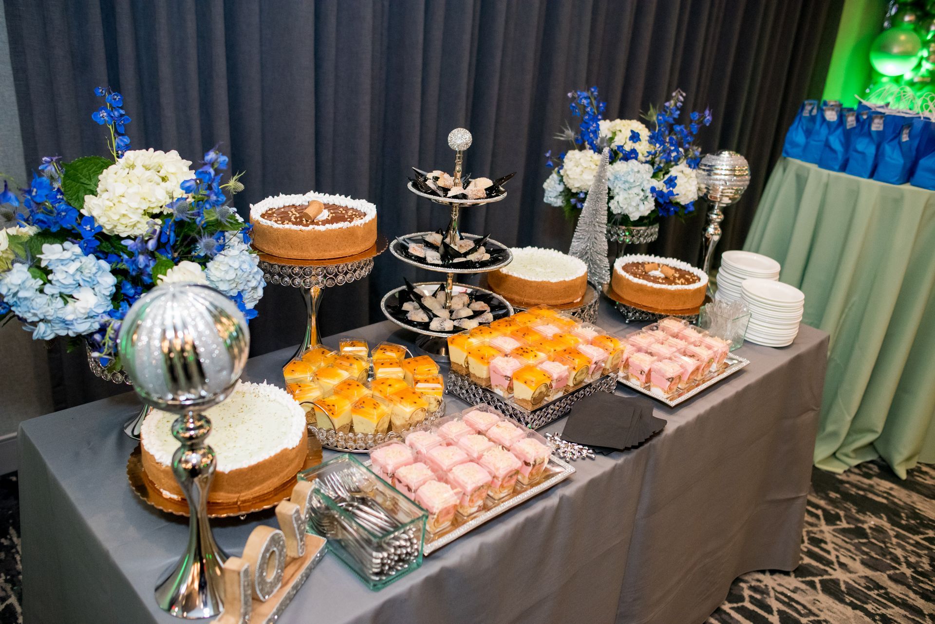 Dessert table with cakes, pastries, and flowers. Blue, gold, and pink treats on a grey table.