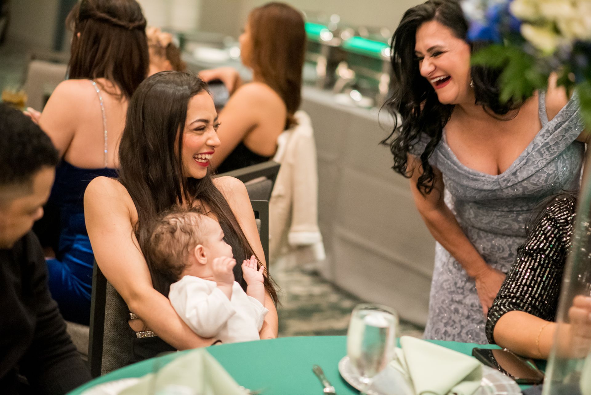 Group of women laughing at an event; one holds a baby. Green table with a floral centerpiece.