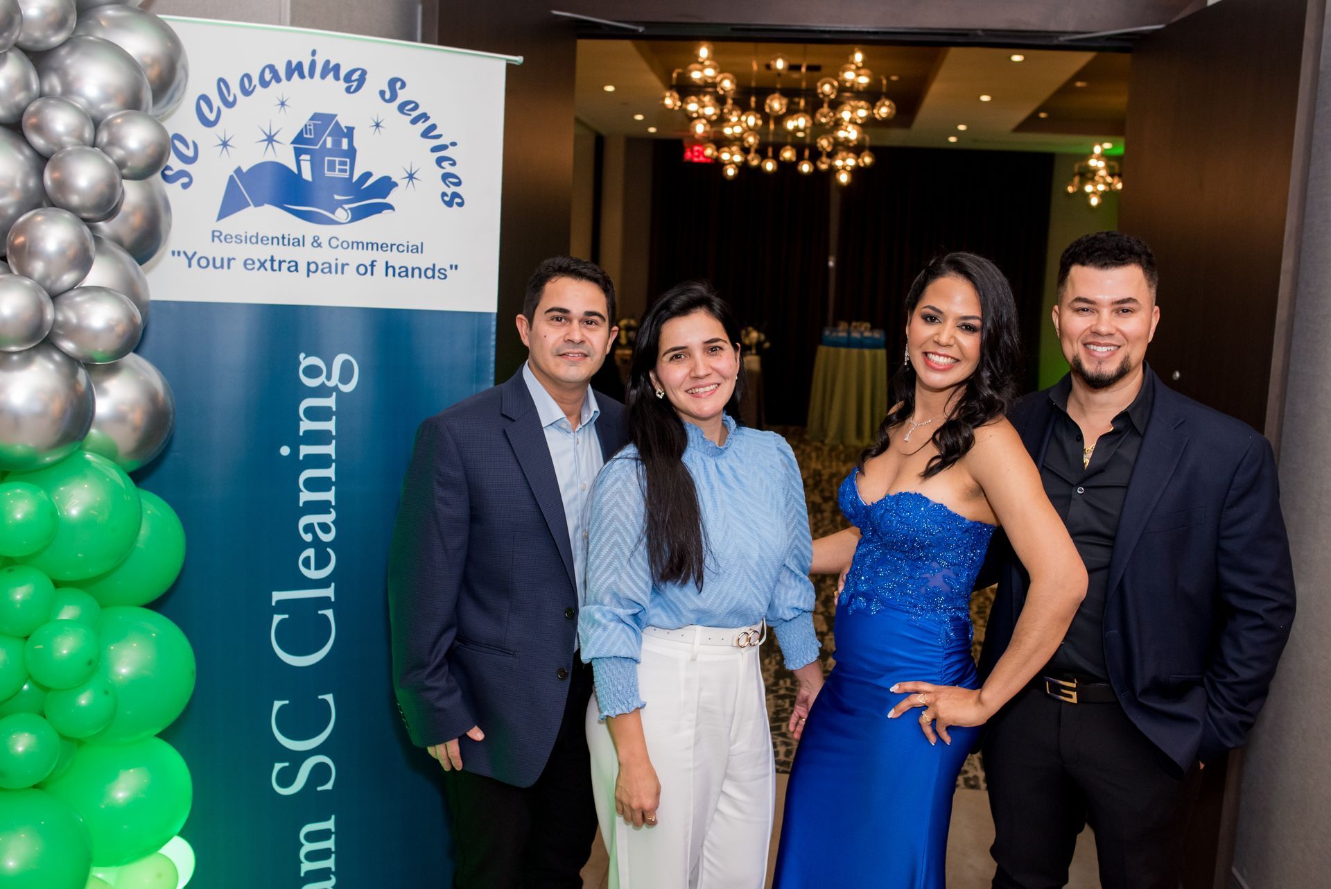 Four people pose by a banner for SC Cleaning Services; balloons, blue dress.