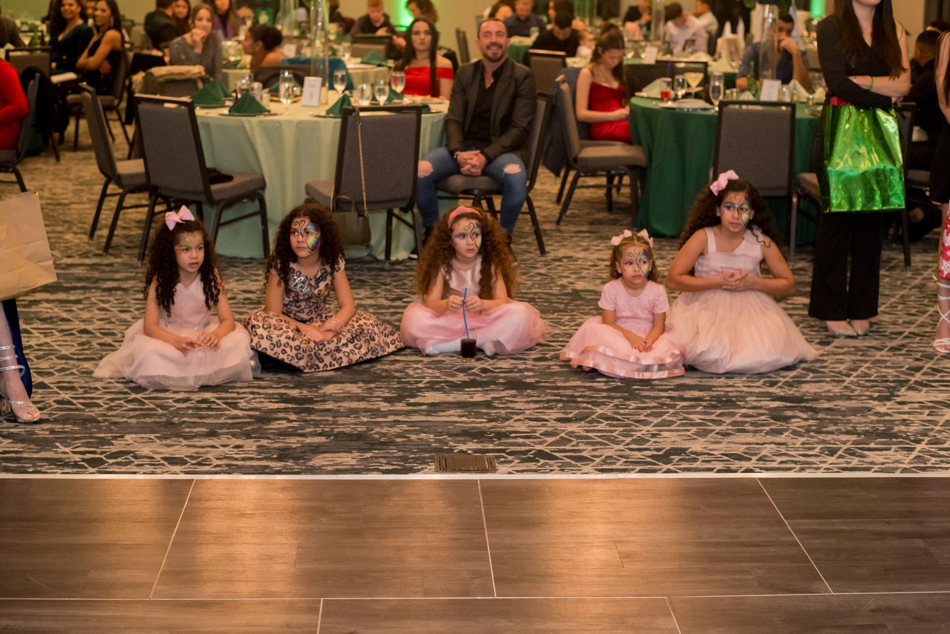 Five young girls in dresses sit on the floor in front of a seated man at an event.