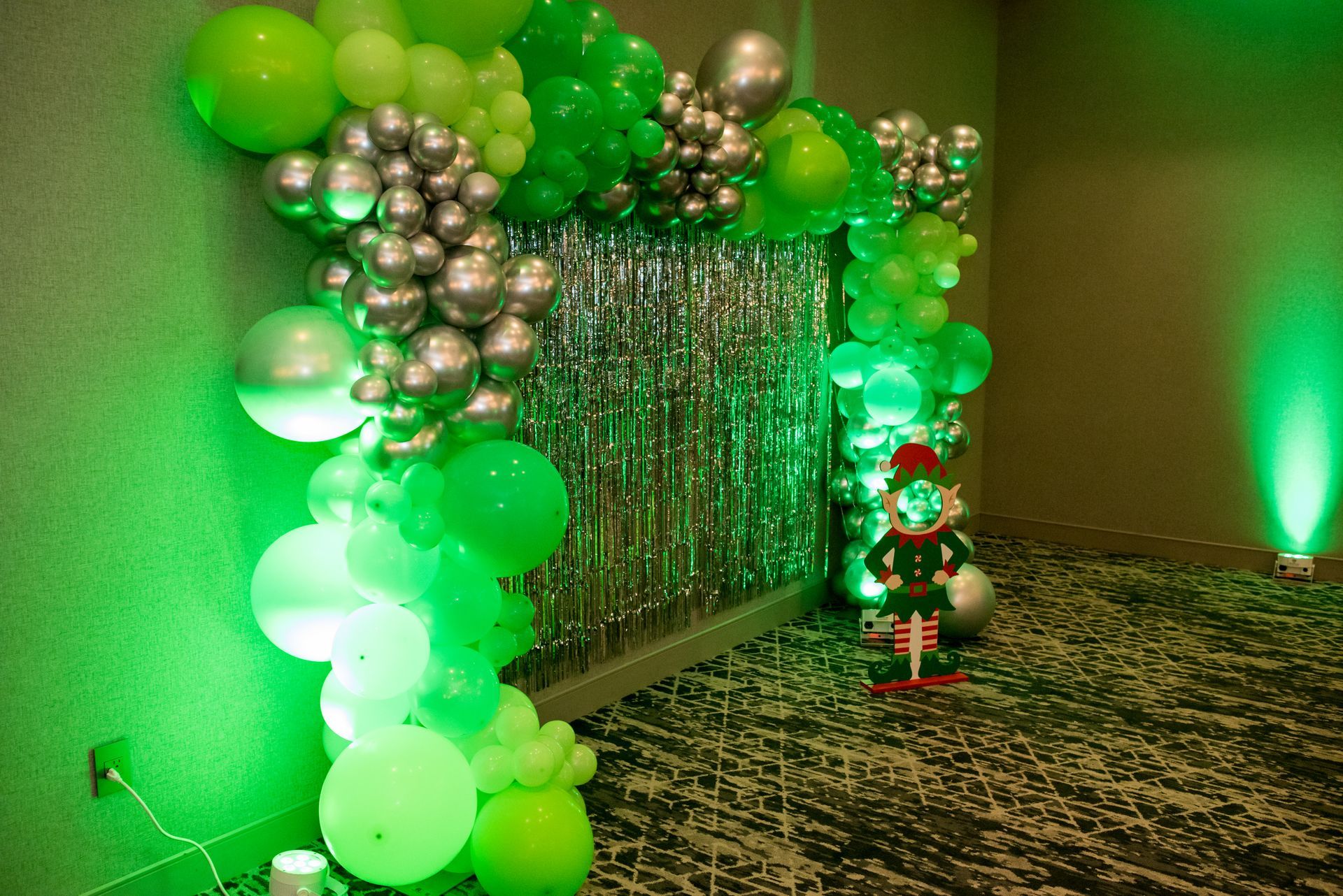 Green and silver balloon arch with a silver curtain backdrop and a holiday elf.