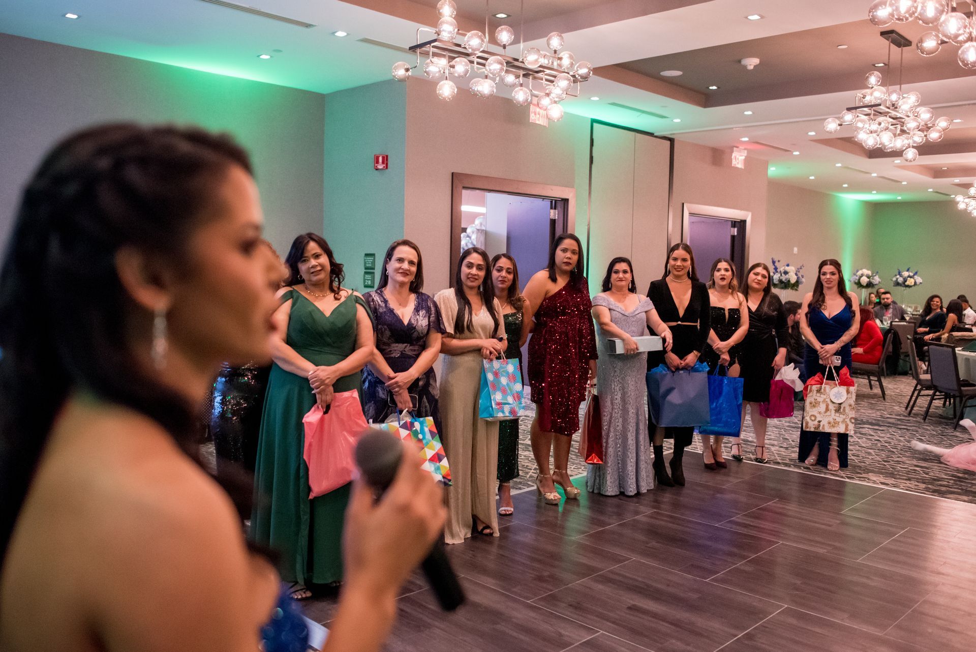 Woman with a microphone addresses a group of women holding gifts in a well-lit event space.