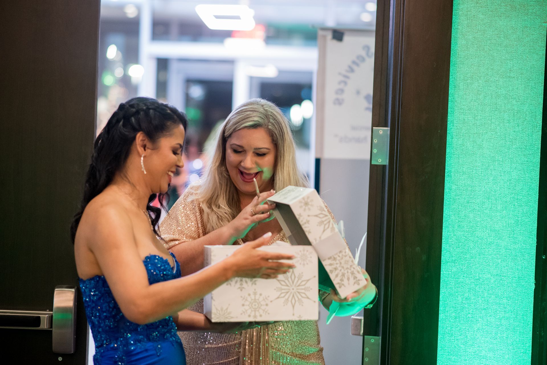 Two women opening a gift box at an event, one in a blue dress, one in a gold sequin dress.
