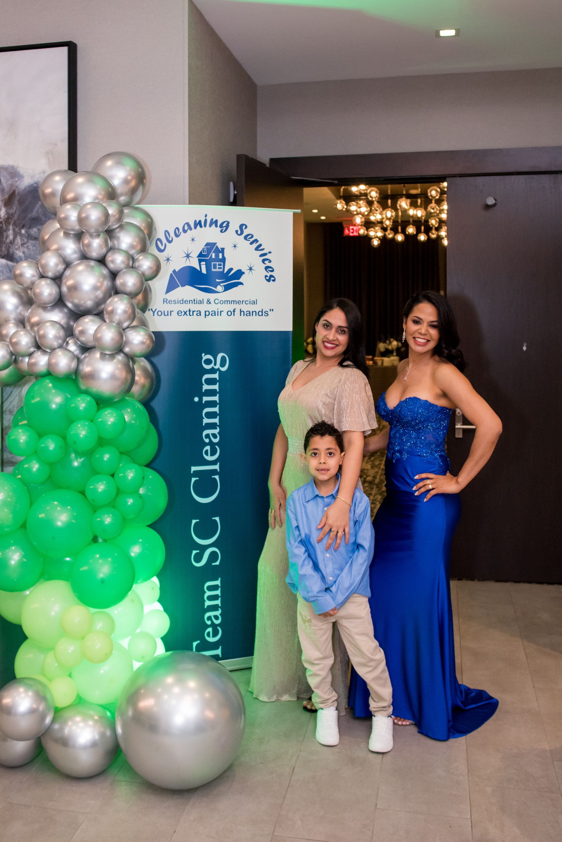 Three people posing near a banner and balloons: two women and a young boy. Women wear formal gowns, boy in casual clothes.