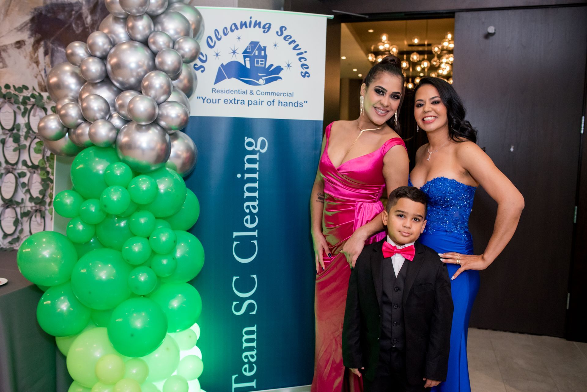 Three people pose by a banner for SC Cleaning: two women in gowns and a boy in a suit by balloons at an event.