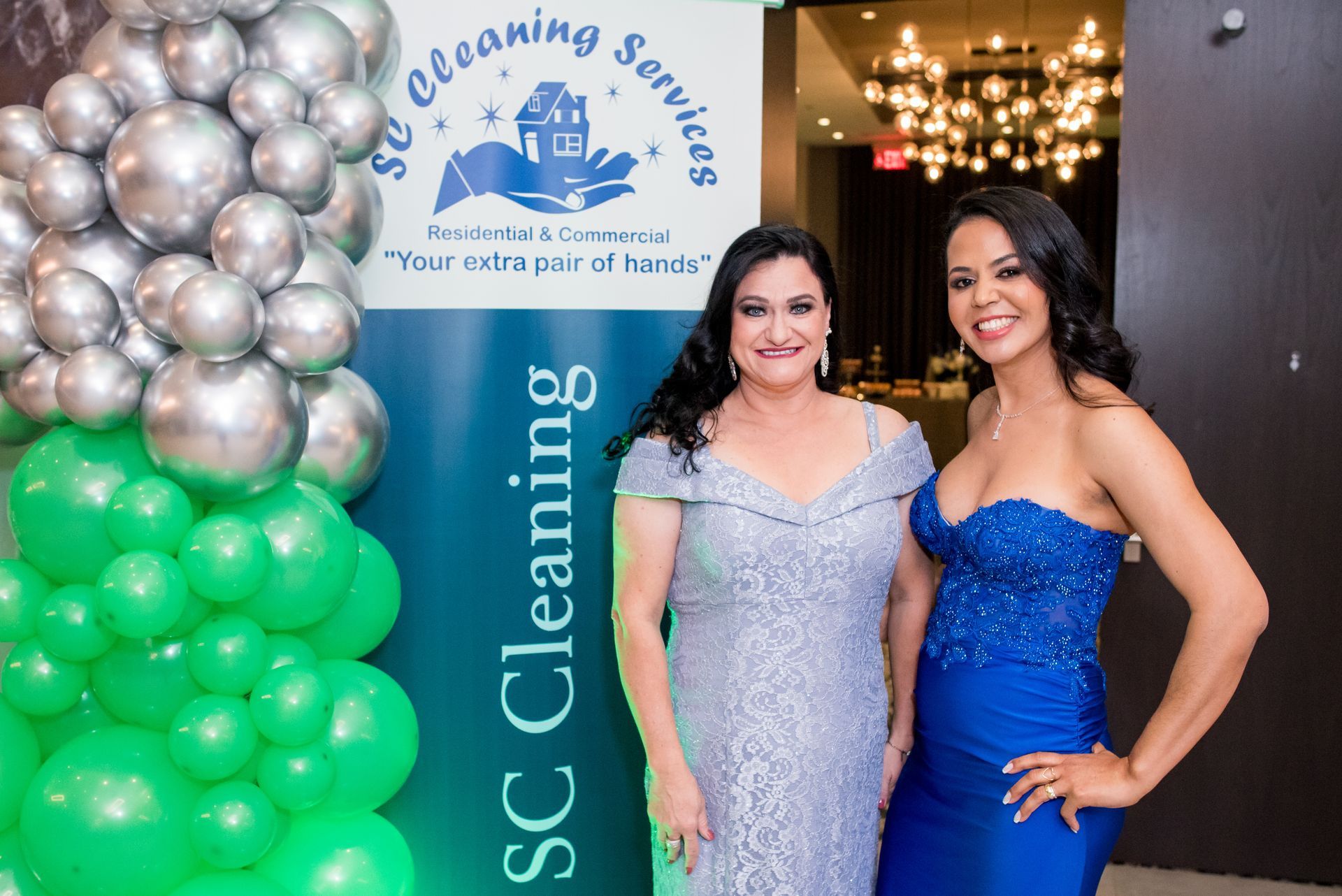 Two women in formal gowns pose near a banner for