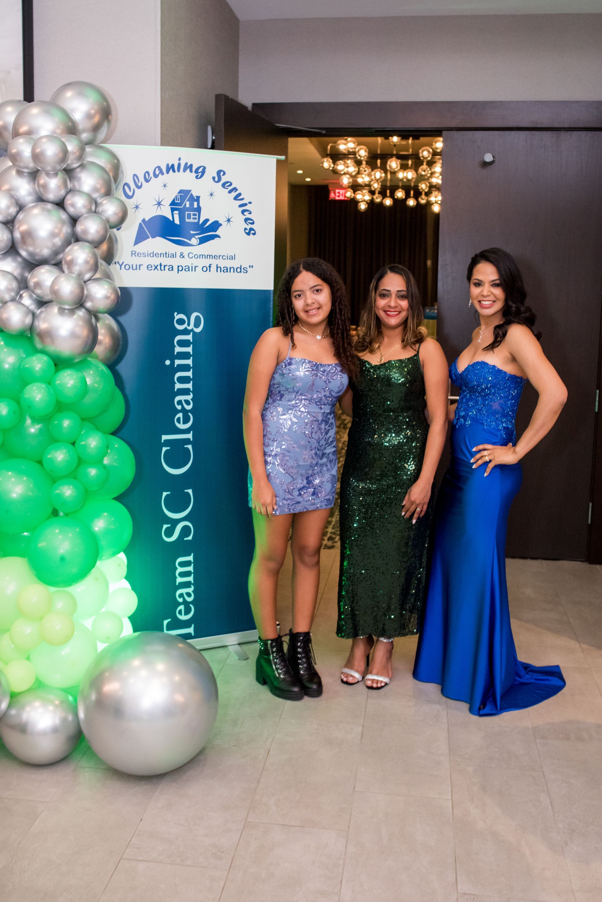 Three women in formal gowns pose by a company banner and balloon arch.