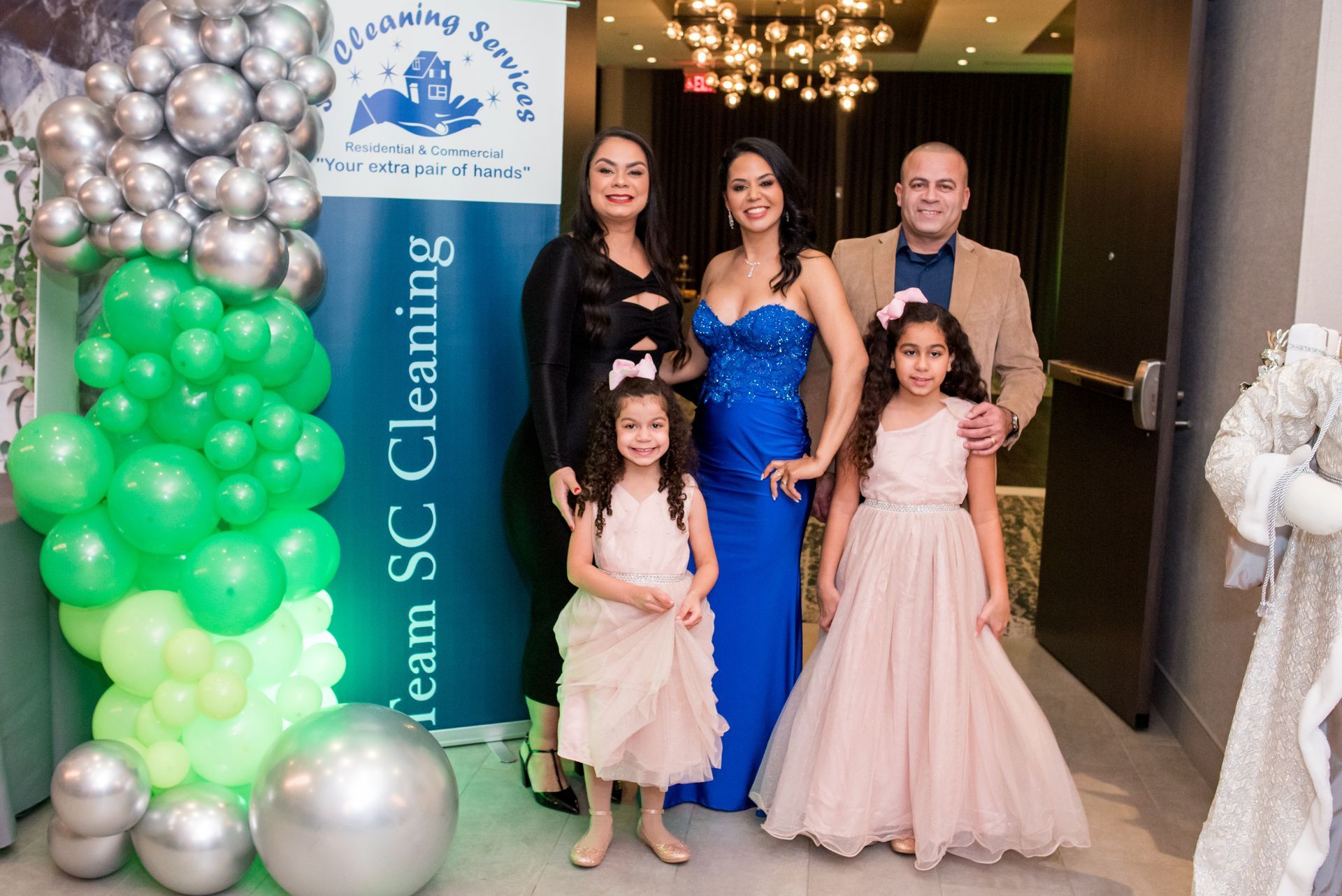 Family posing at event, smiling. Woman in blue gown, green and silver balloons, banner for cleaning service.