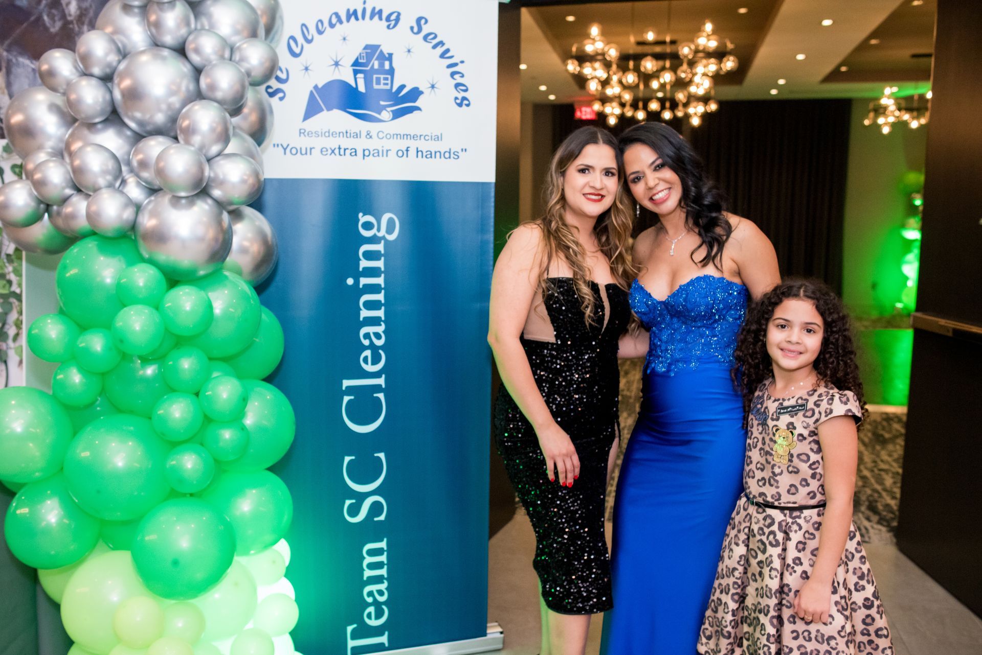 Three people pose by a banner with balloons. Woman in blue dress, black dress, and a girl in leopard print dress.