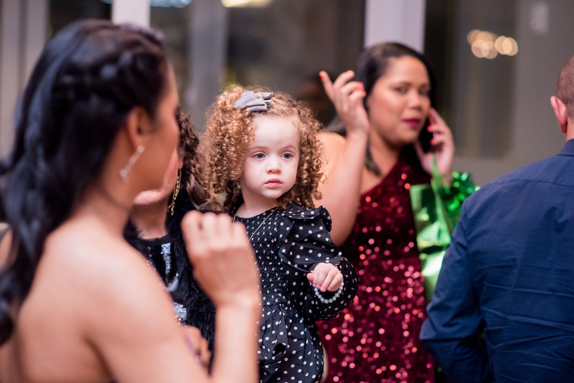 Child with curly hair in black polka dot dress, surrounded by adults at a party.