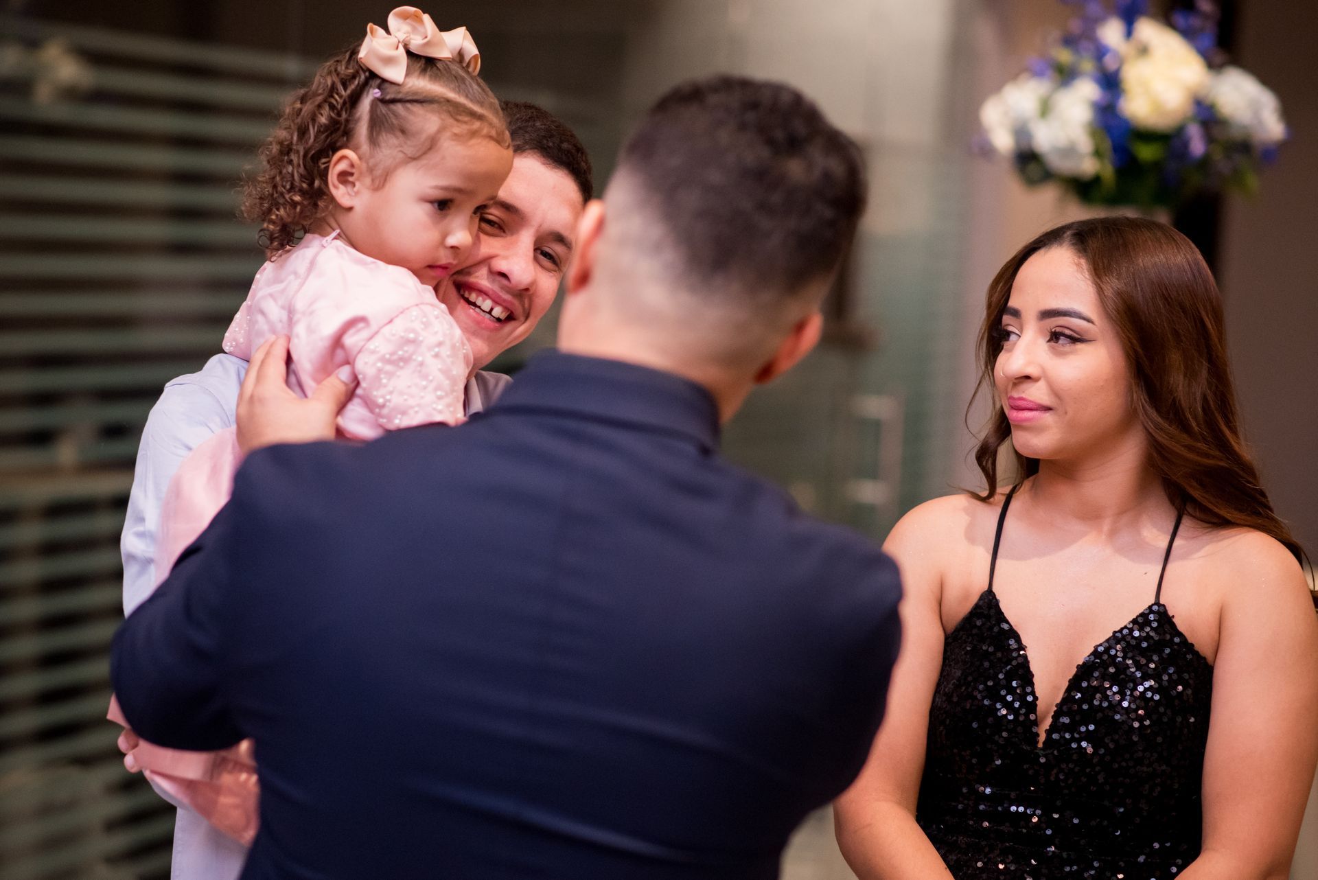 Man holding child, smiling at another man. Woman in sequined dress smiles. Formal setting.