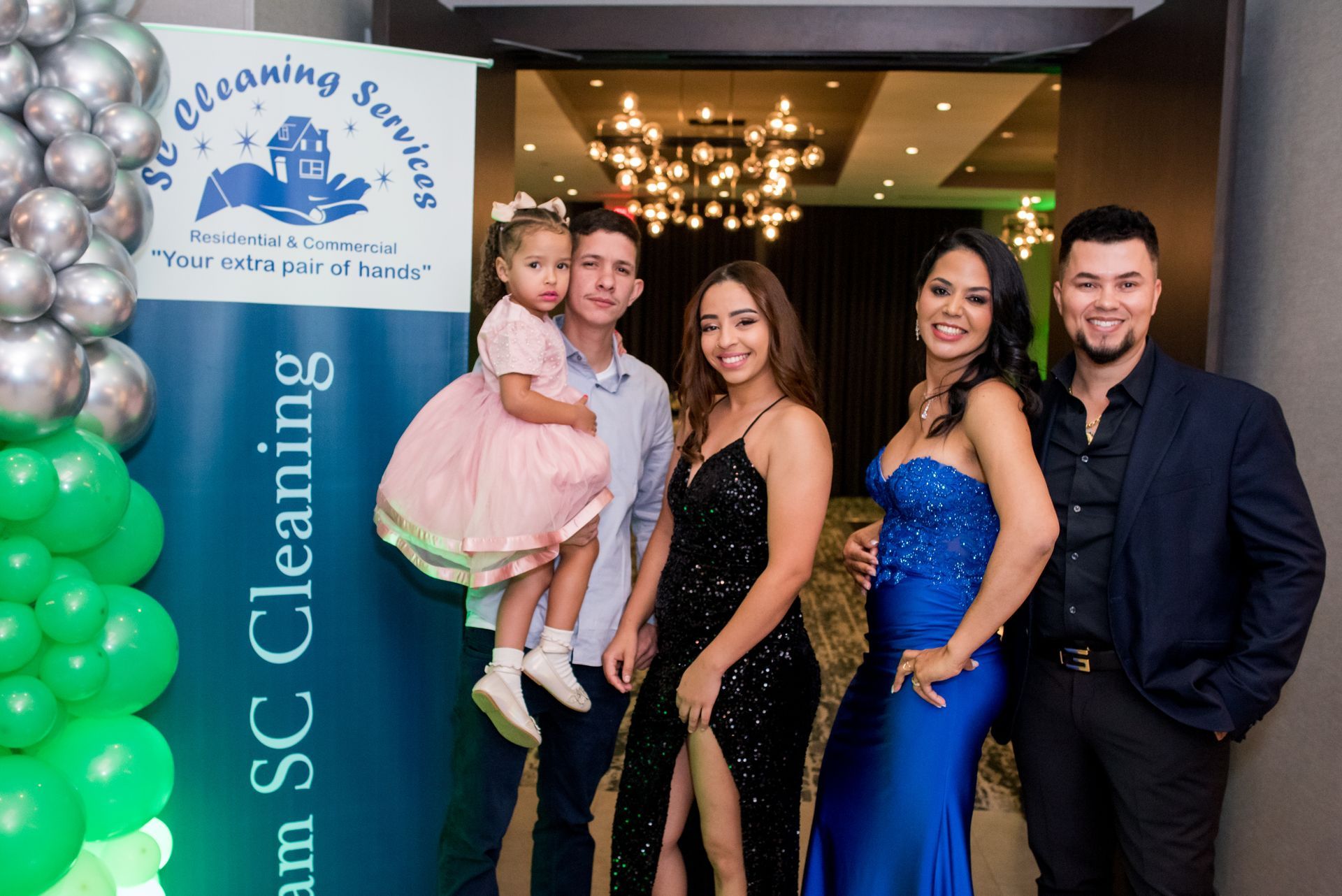 Group of five people posing near an SC Cleaning Services banner, balloons, and chandelier; formal attire.