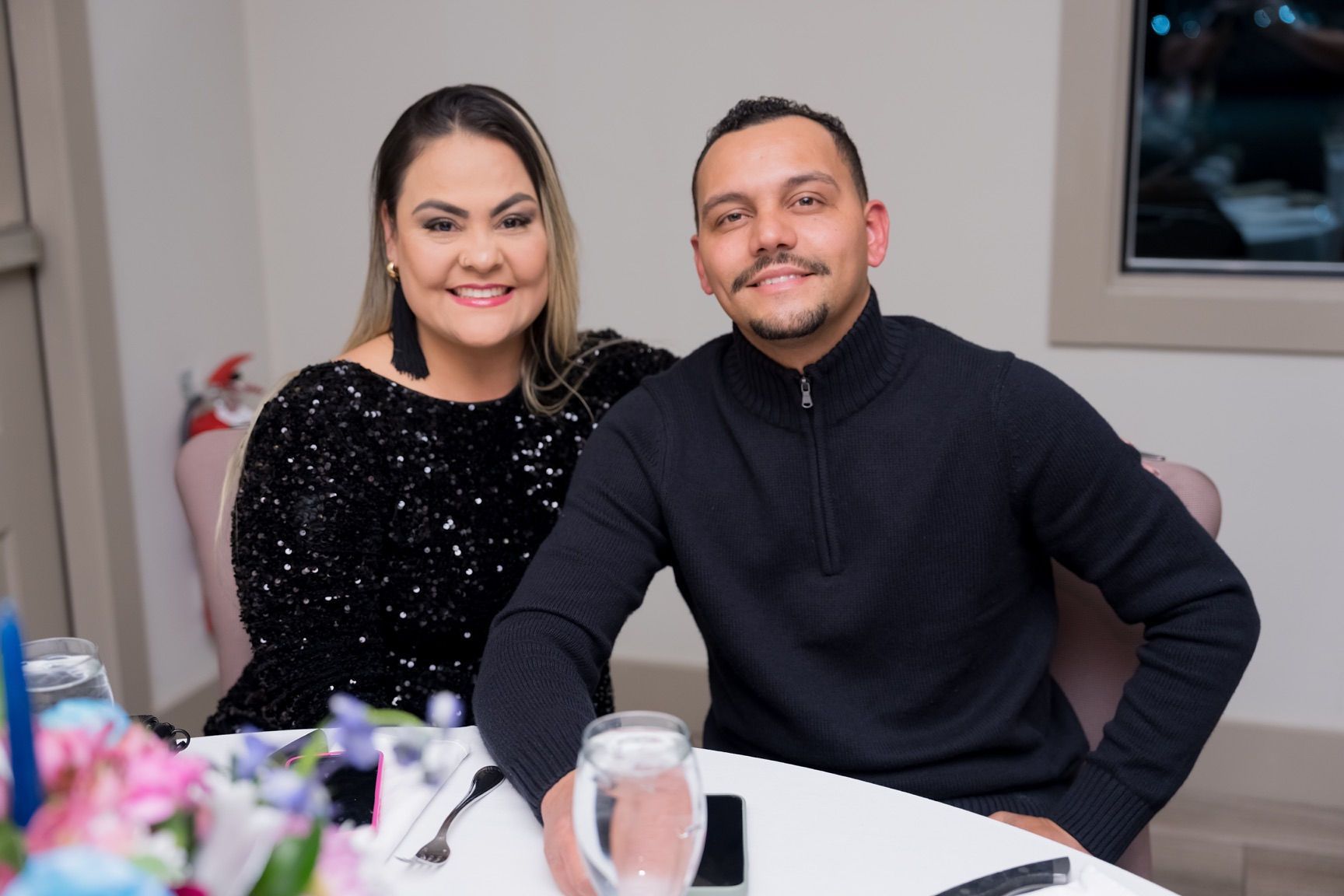 Woman with dark hair in a black sequin top and man in a black sweater smiling at a table.
