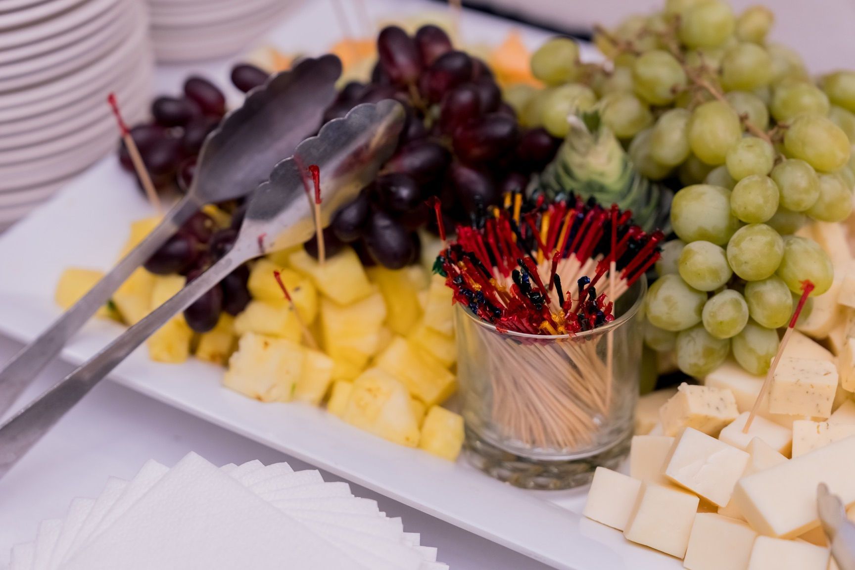 A platter of fruit (grapes, pineapple) and cheese, with tongs and toothpicks.