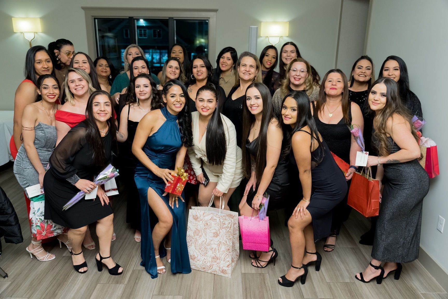 Group of women in formal attire posing in a room, holding gifts, smiling.