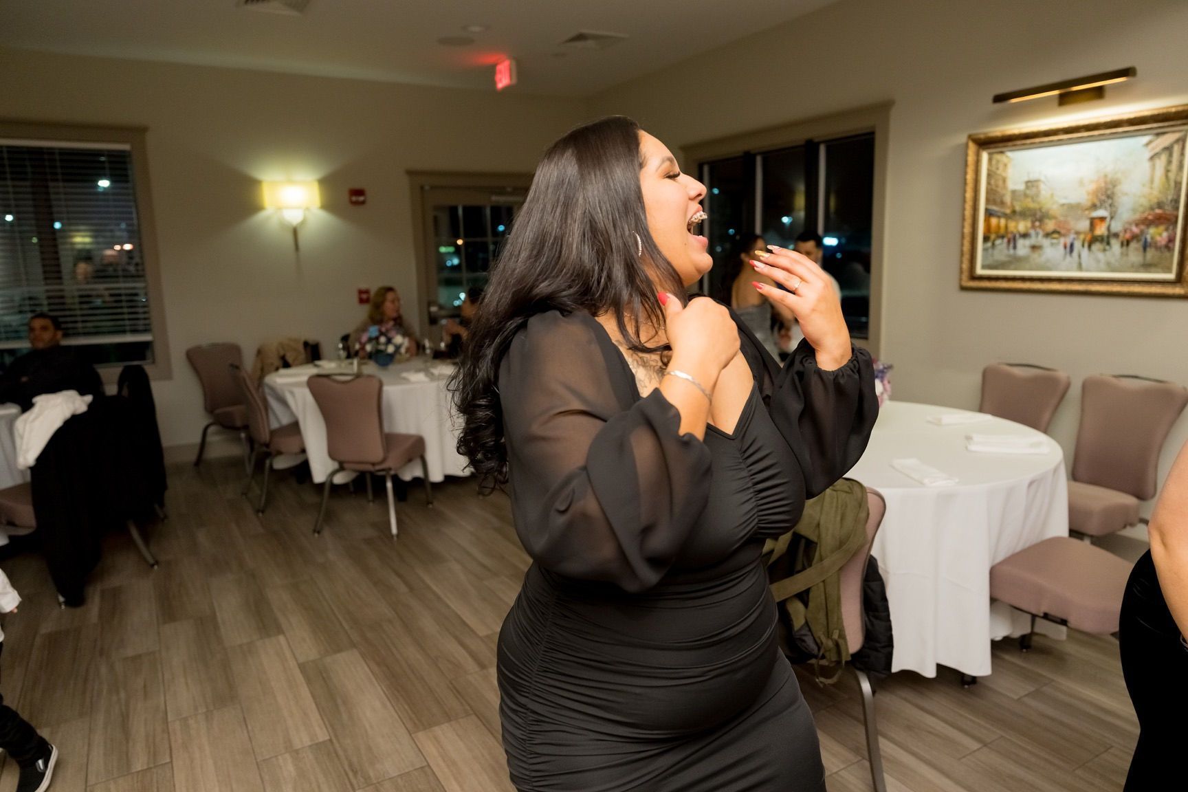 Woman in black dress laughing, dancing in a party setting with round tables and artwork.