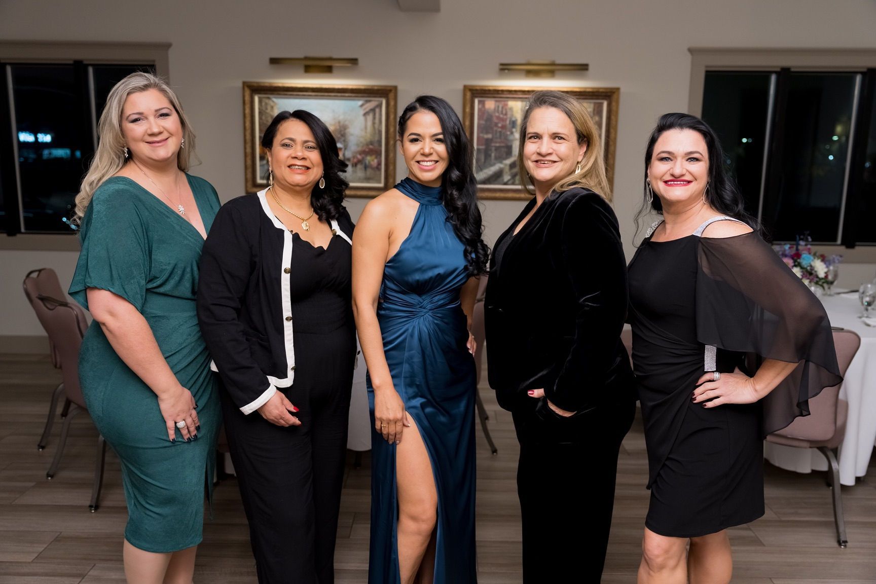 Five women in formal attire pose indoors with paintings in the background.