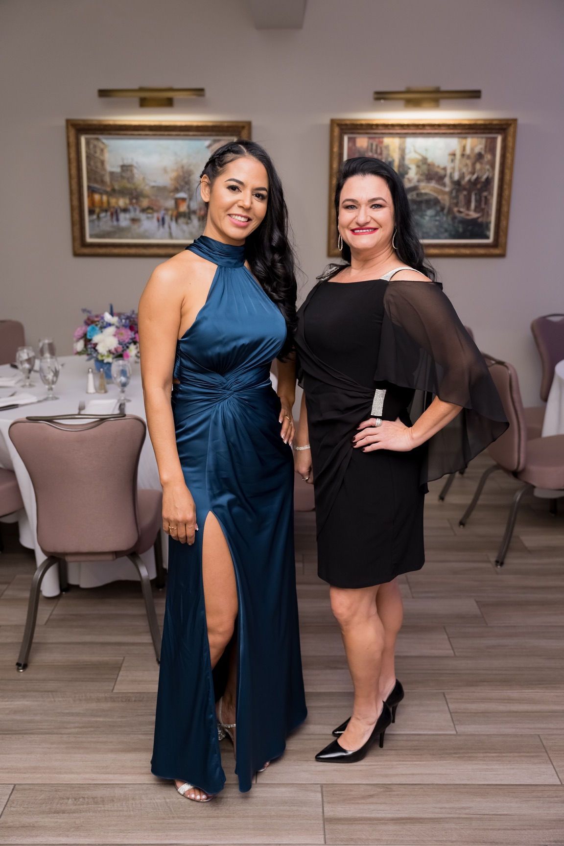 Two women in formal wear pose indoors; one in a teal gown with a high slit, the other in a black dress.