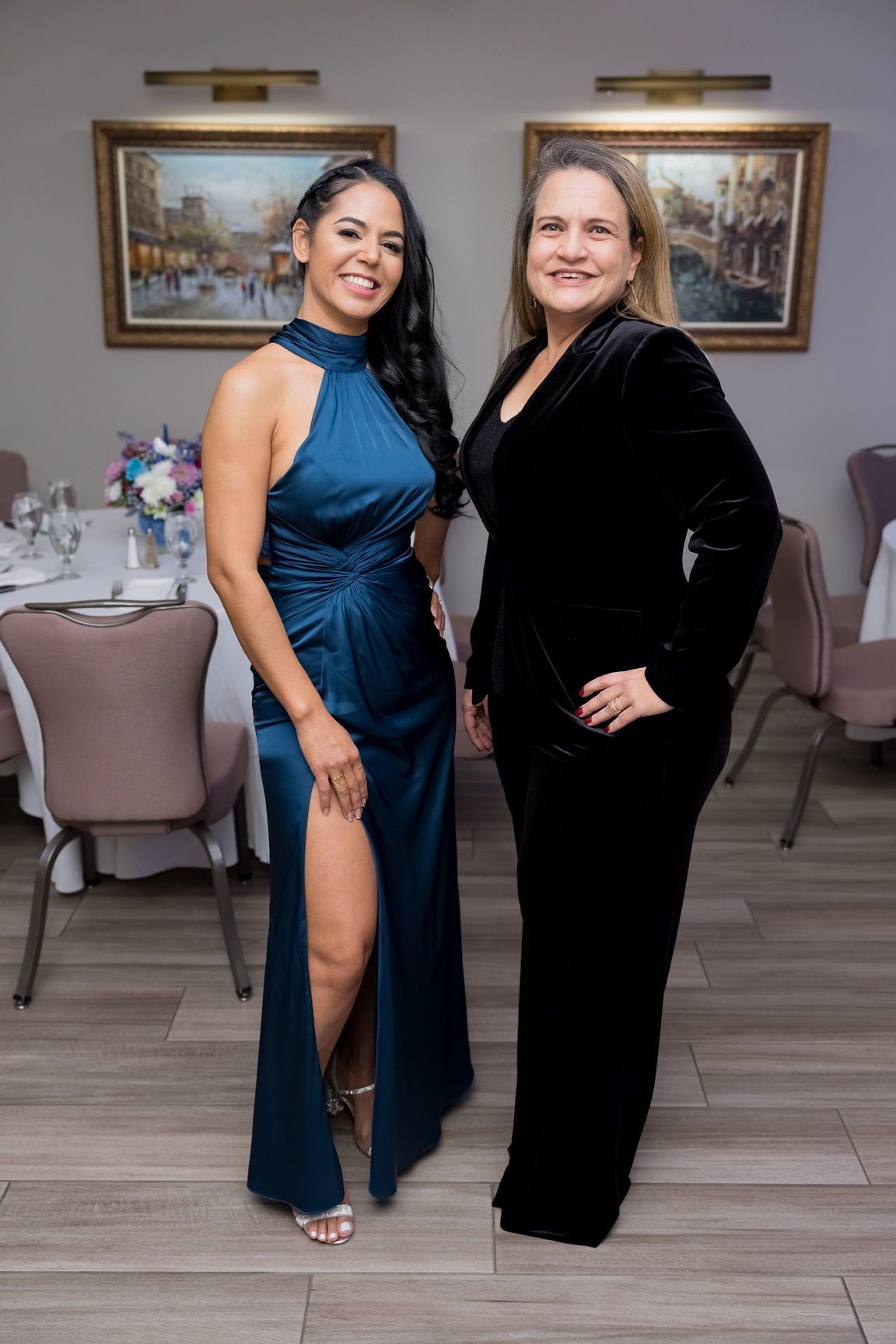 Two women pose for photo at an event. One in teal dress with slit, the other in black suit.