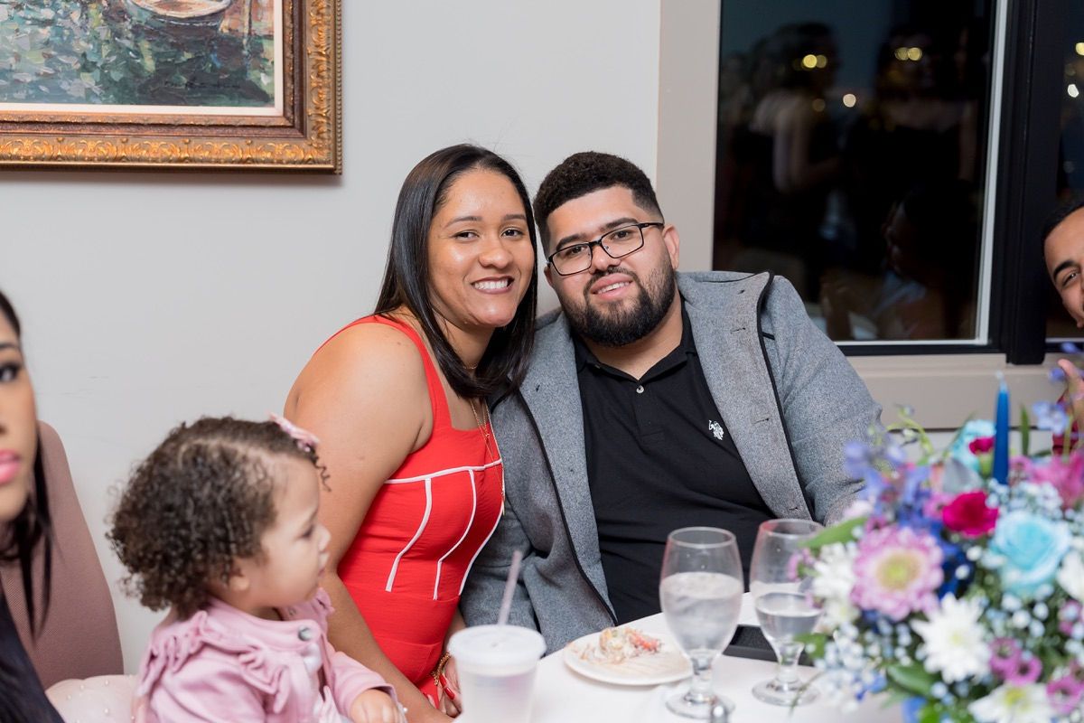 Family at a table: woman in red dress and man in glasses smile. Child in pink jacket sits nearby.