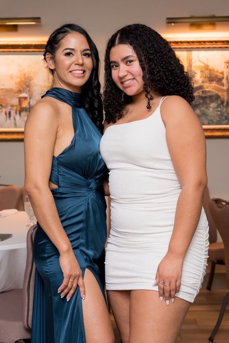 Two women in formal attire pose smiling in front of a painting at a formal event. One in blue, one in white.