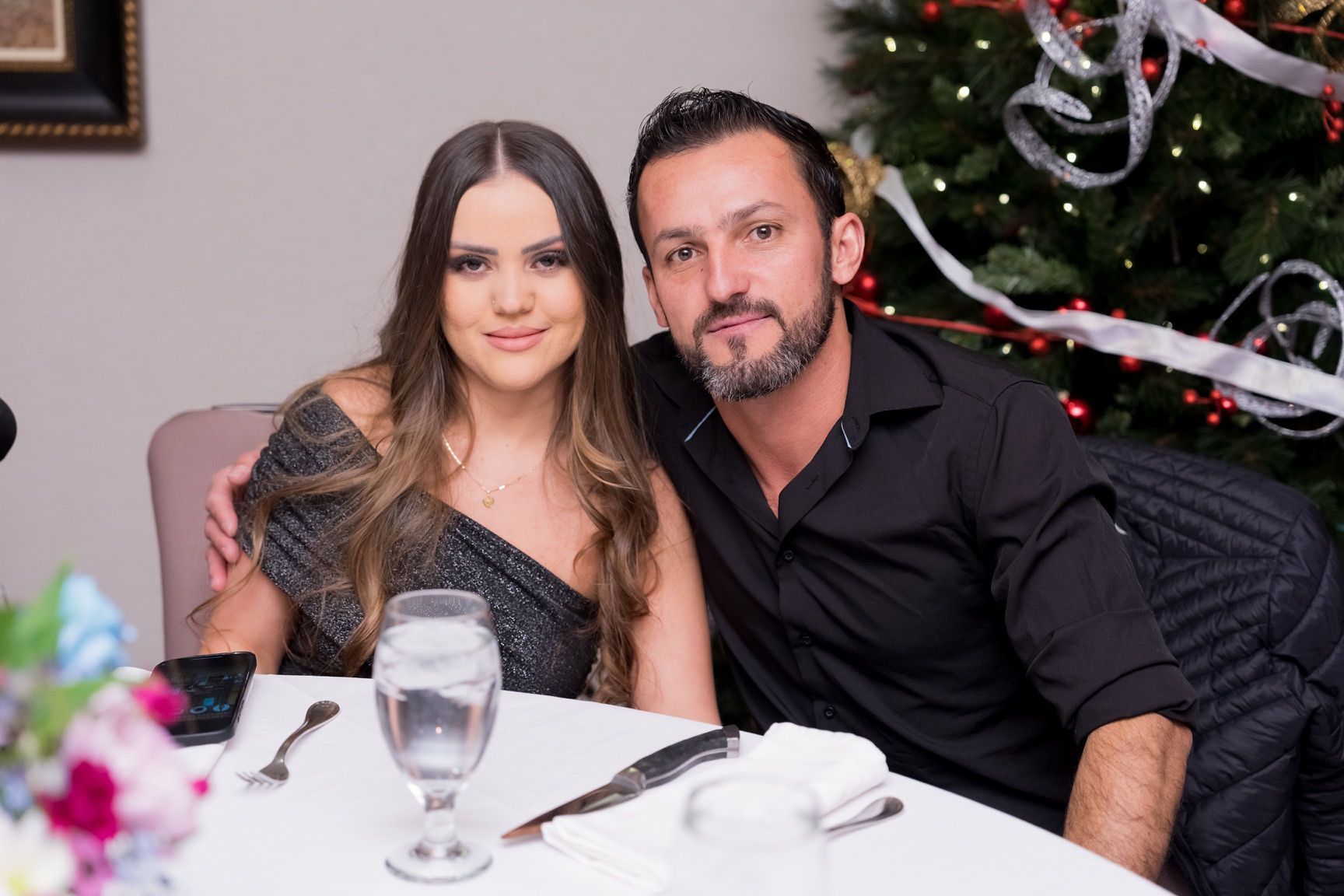 Woman and man smiling at table, by a Christmas tree. She wears a sparkly dress. He wears a black shirt.