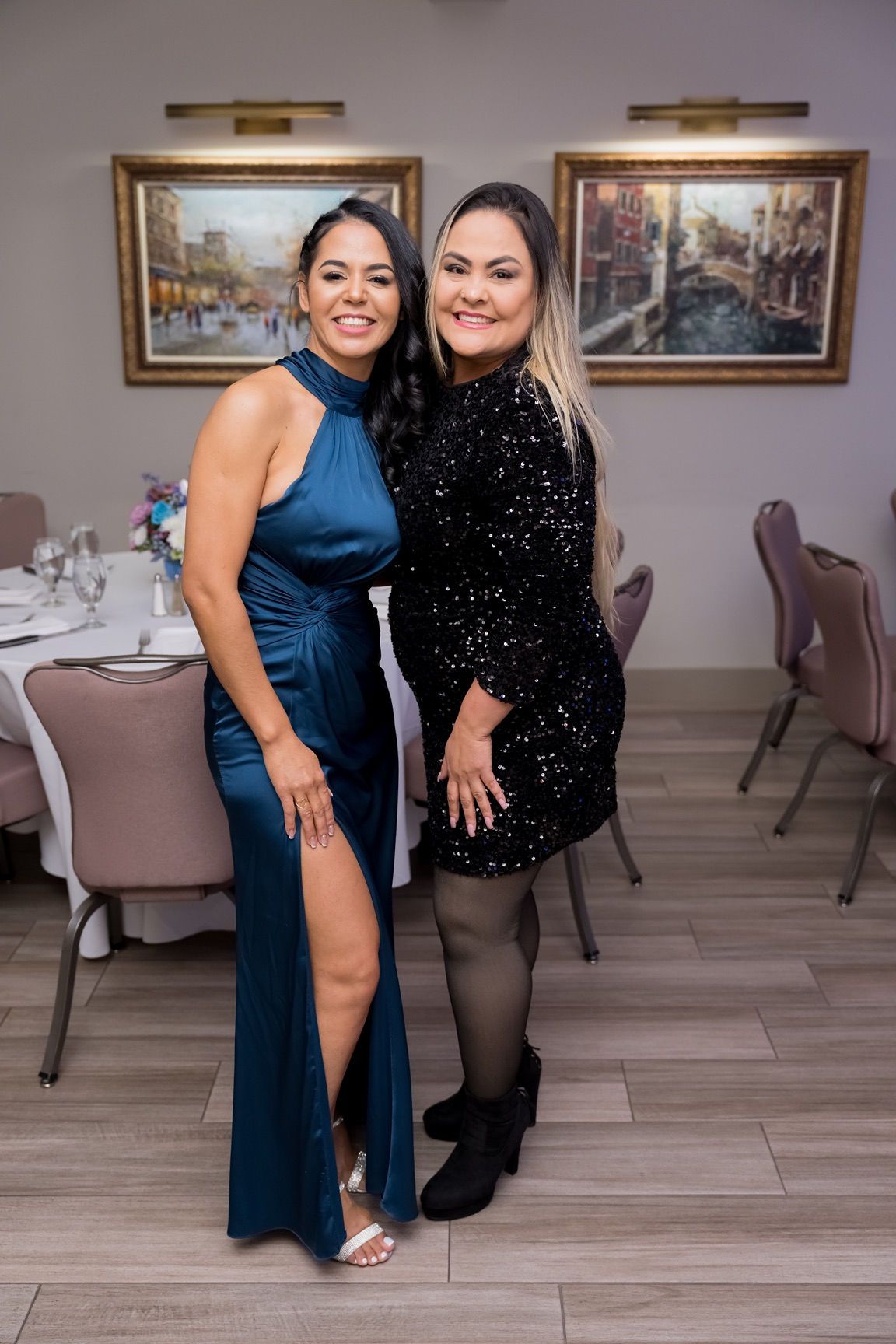 Two women smiling, posing for photo at event. One wears blue gown, other wears black sequin dress, indoor setting.