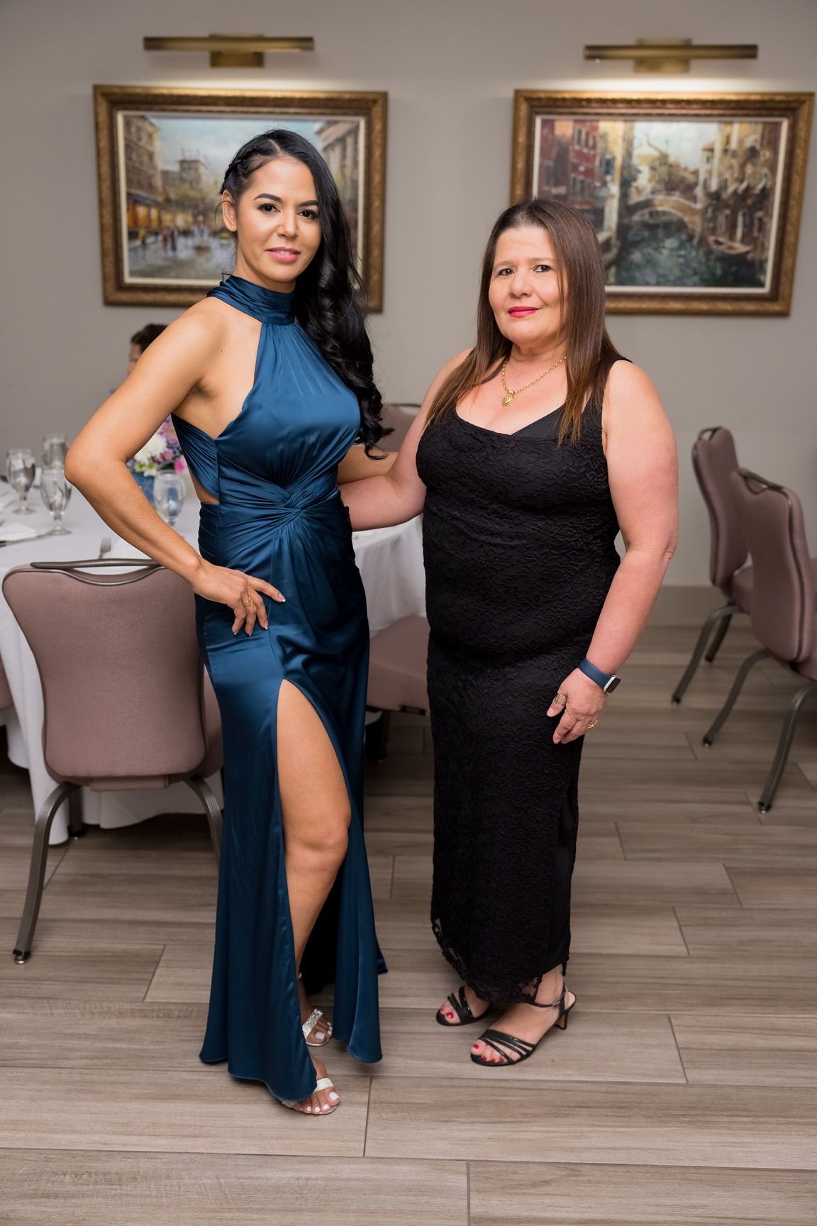 Two women pose in a formal setting. One wears a blue dress with a high slit, the other a black dress.