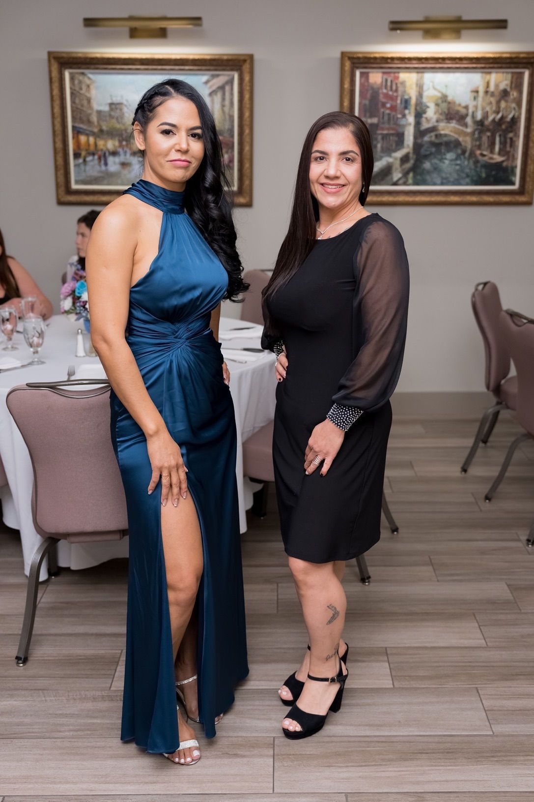Two women at a formal event. One in a blue slit dress, the other in a black dress.