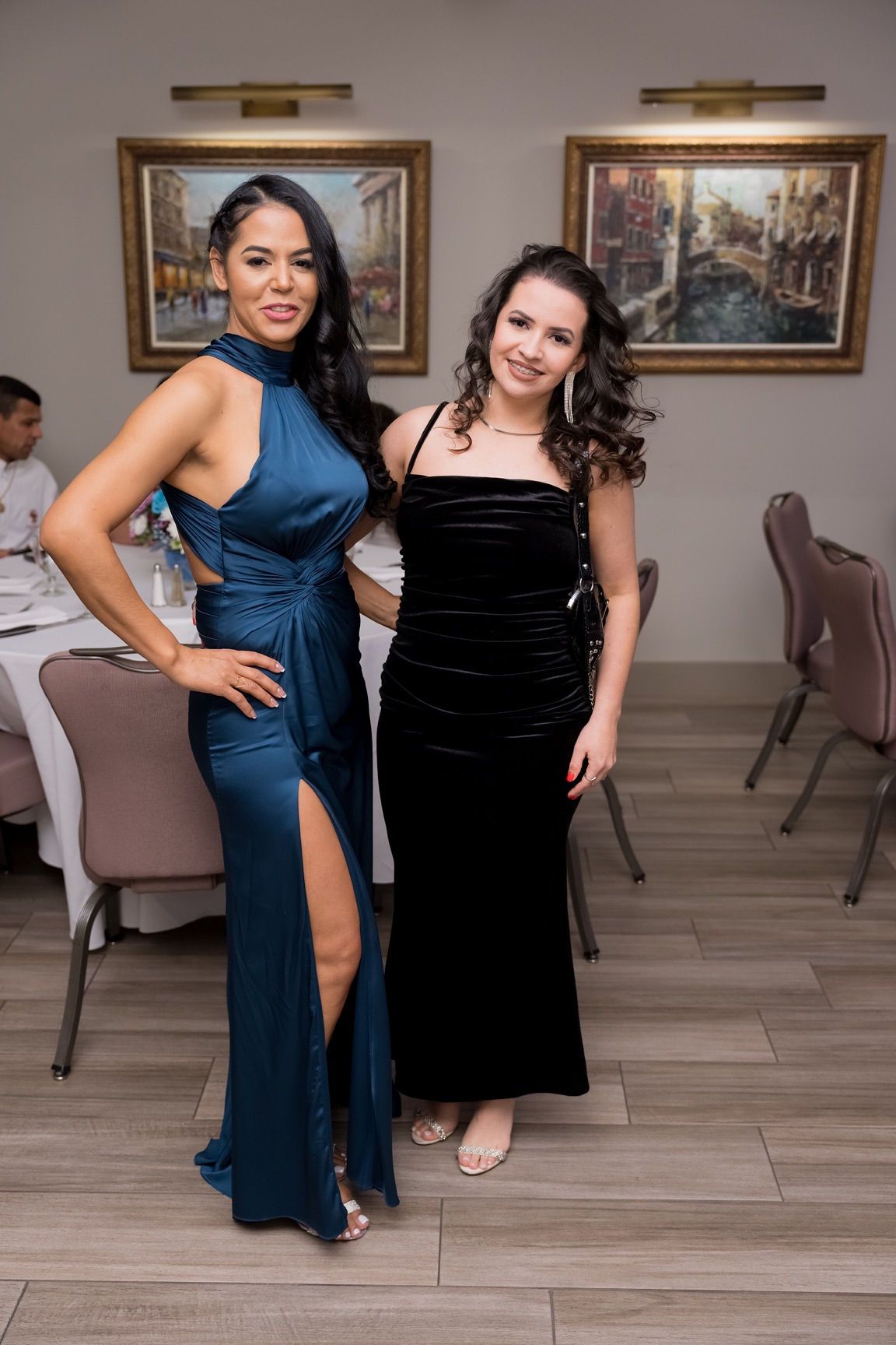 Two women in formal dresses pose indoors, one blue, one black.