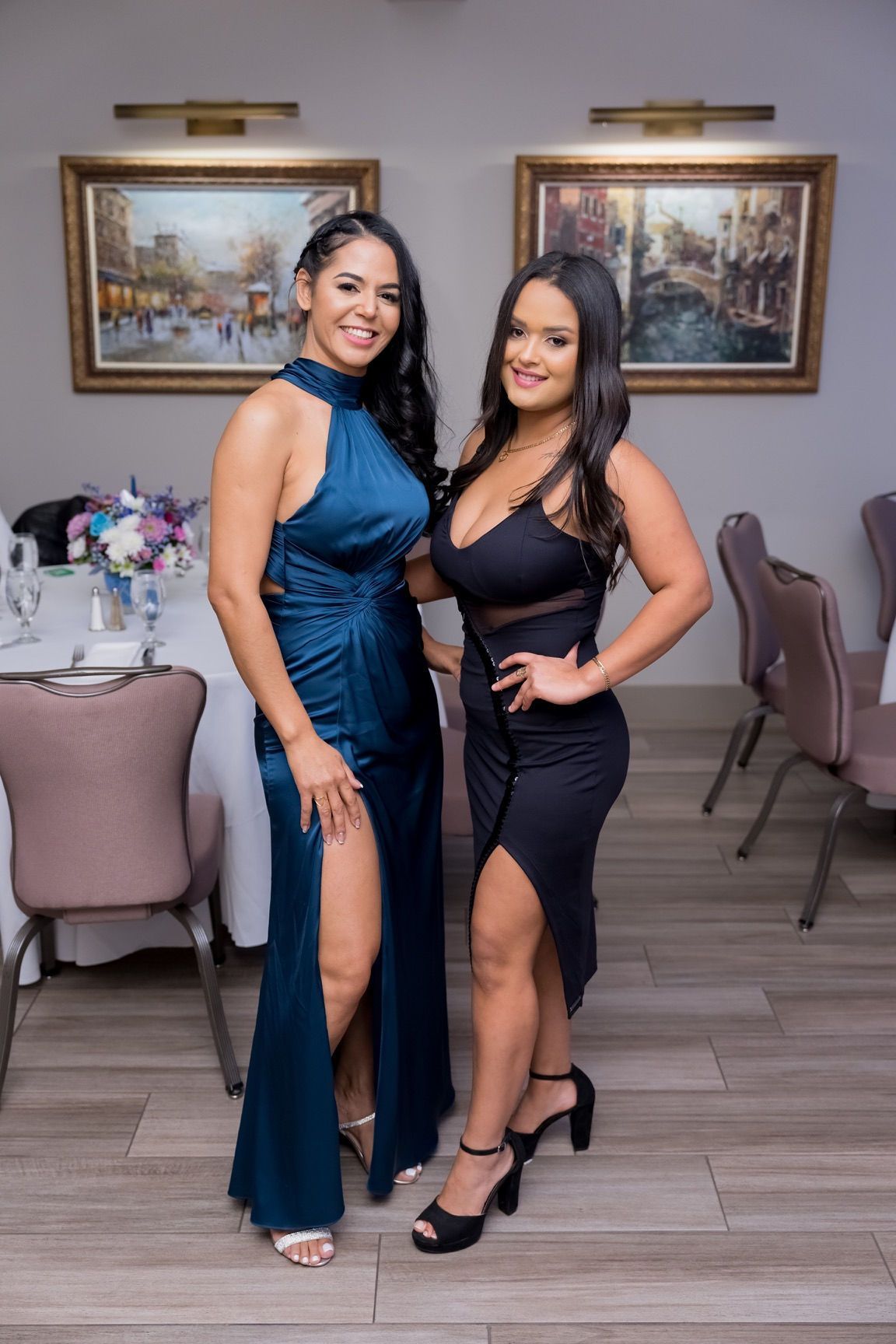 Two women in formal dresses pose indoors, smiling. One in teal, the other in black with cutouts.