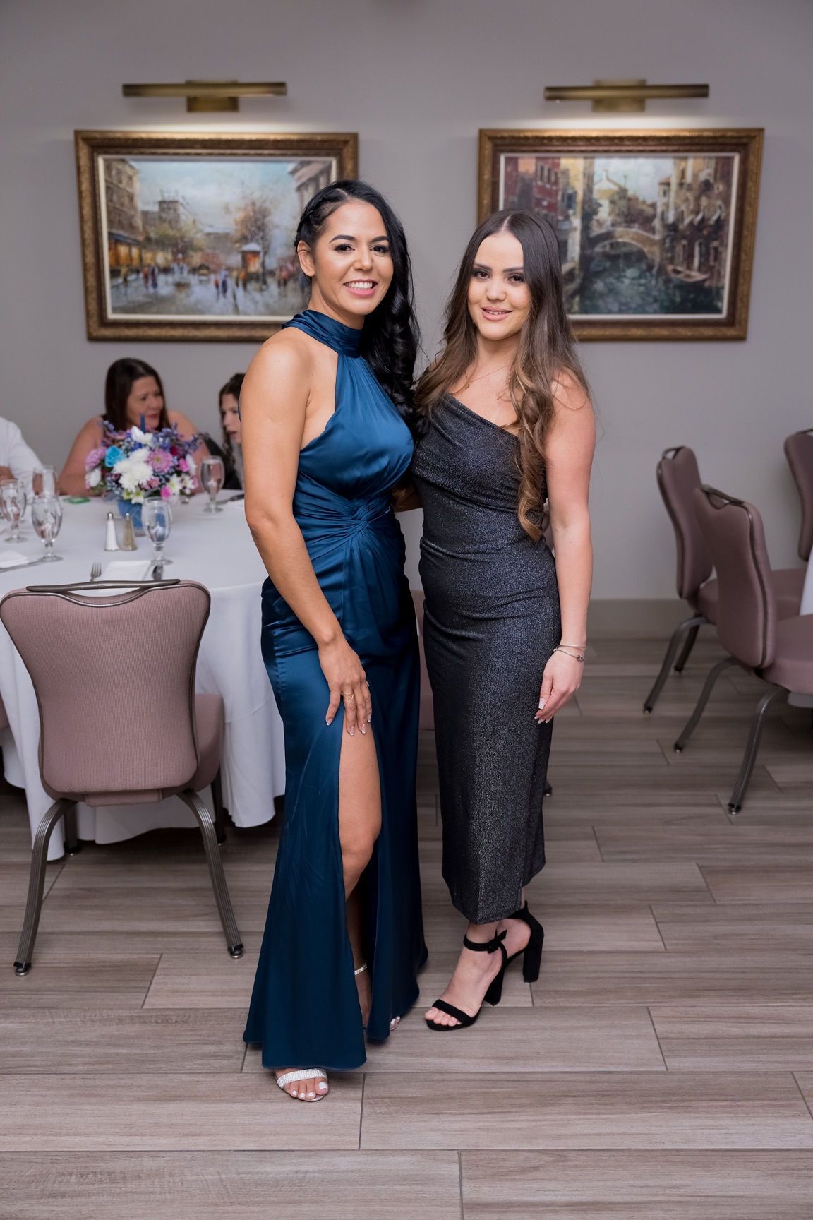 Two women in formal dresses at an event, posing in a room with tables, artwork.