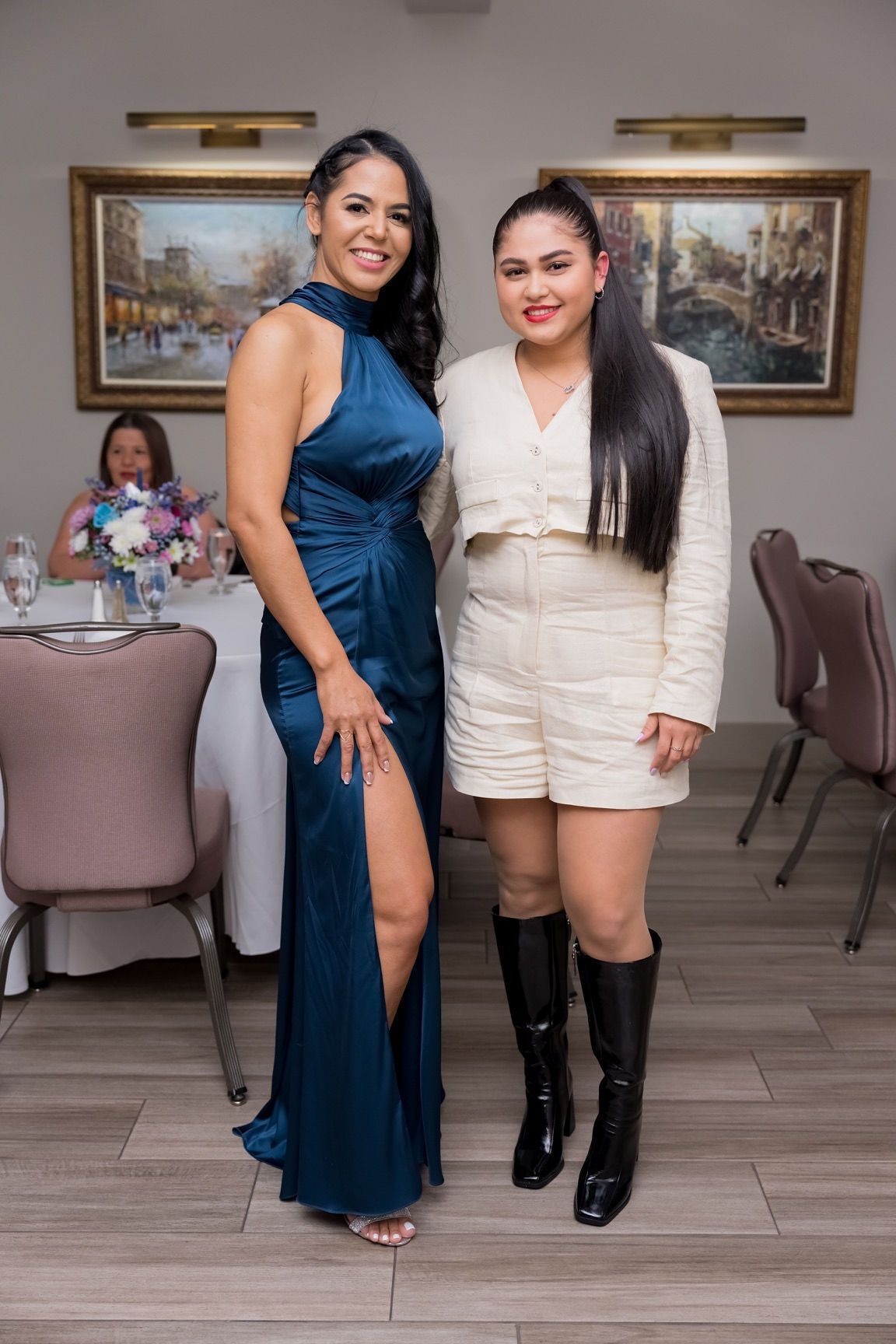 Two women at an event. One in a blue gown with a high slit, the other in a white romper and boots.