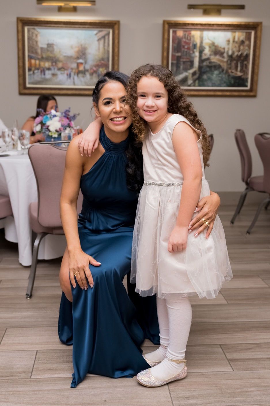 Woman in blue dress and girl in white dress smile, hugging in a room with paintings.