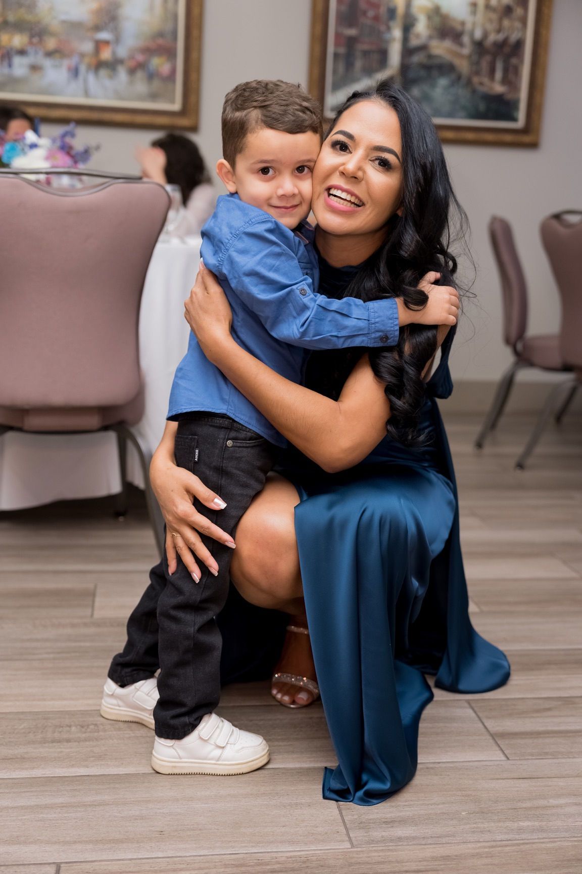 Woman in blue dress and boy in blue shirt embrace, smiling at the camera. Indoors.