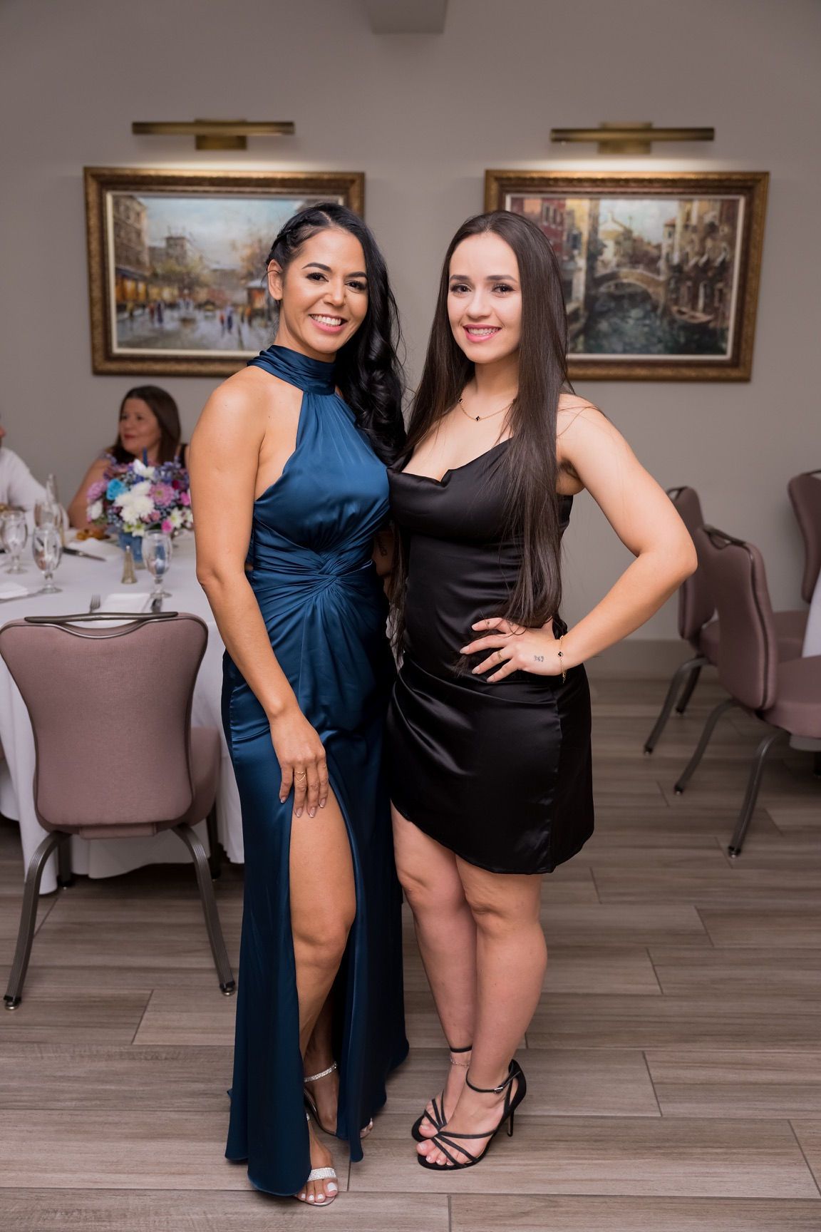 Two women pose at an event. One in a blue gown, the other in a black dress, indoors.