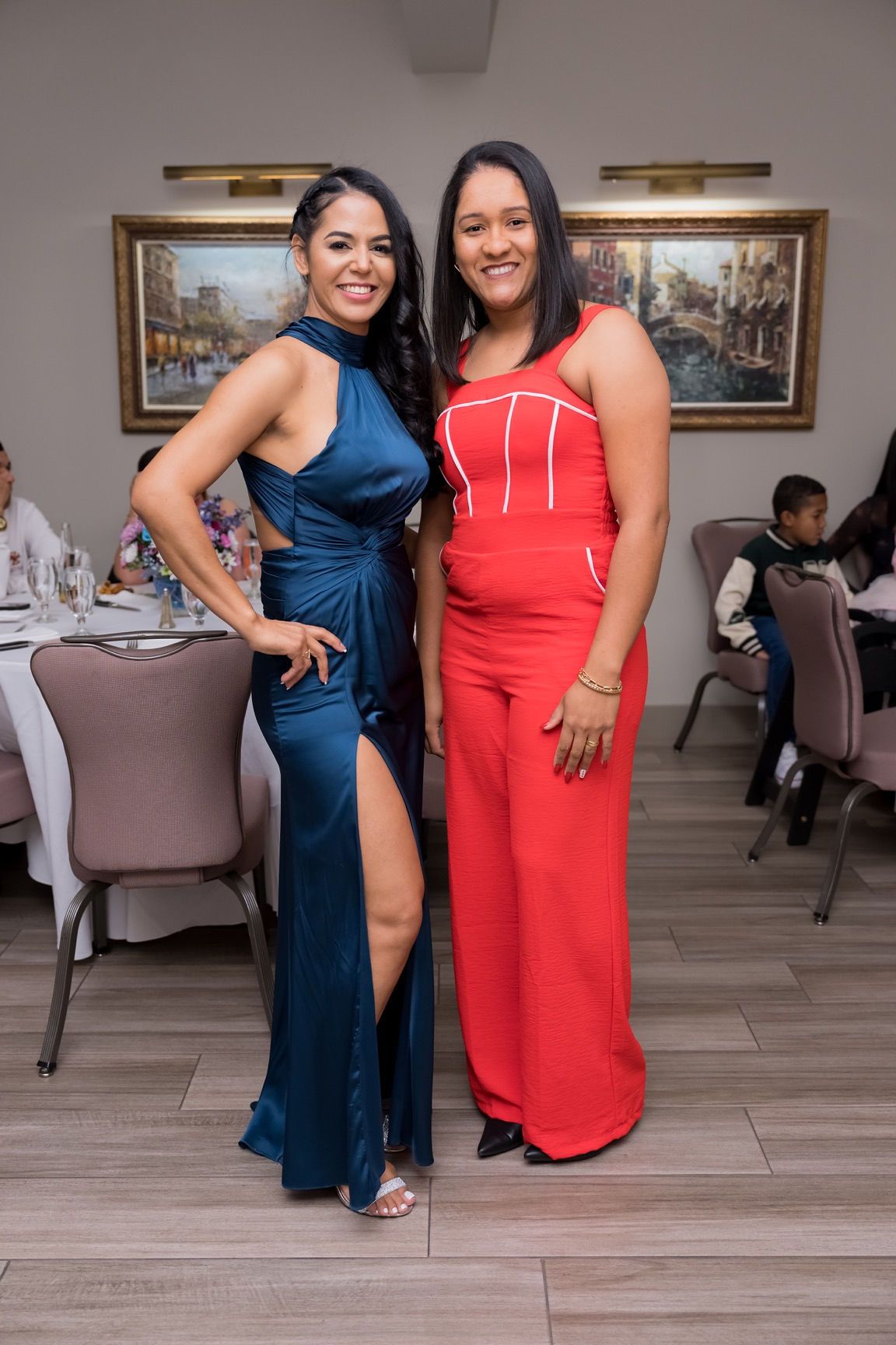 Two women pose at an event. One in blue gown, the other in red jumpsuit. Both smile.