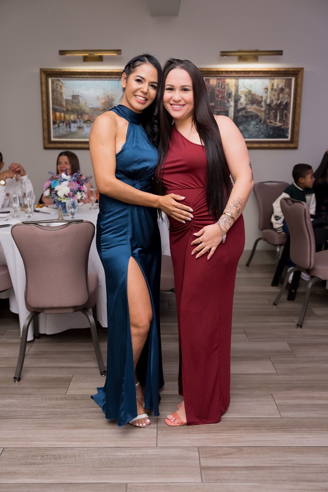 Two women smiling, posing in formal dresses at an event. One wears teal with a high slit, the other burgundy.
