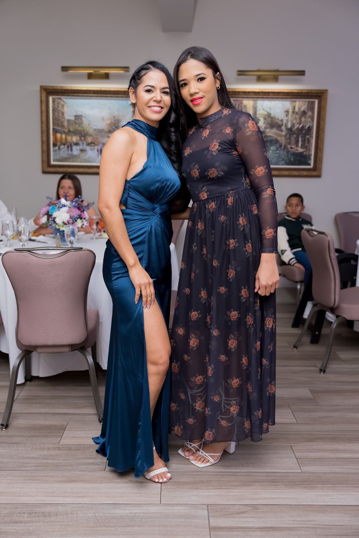 Two women at a formal event, one in a teal dress, the other in a floral gown. Indoors with tables set.