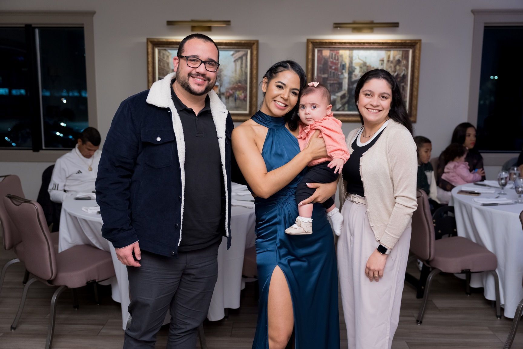 Family posing at a formal event; includes a woman holding a baby, two other adults, and dining tables.