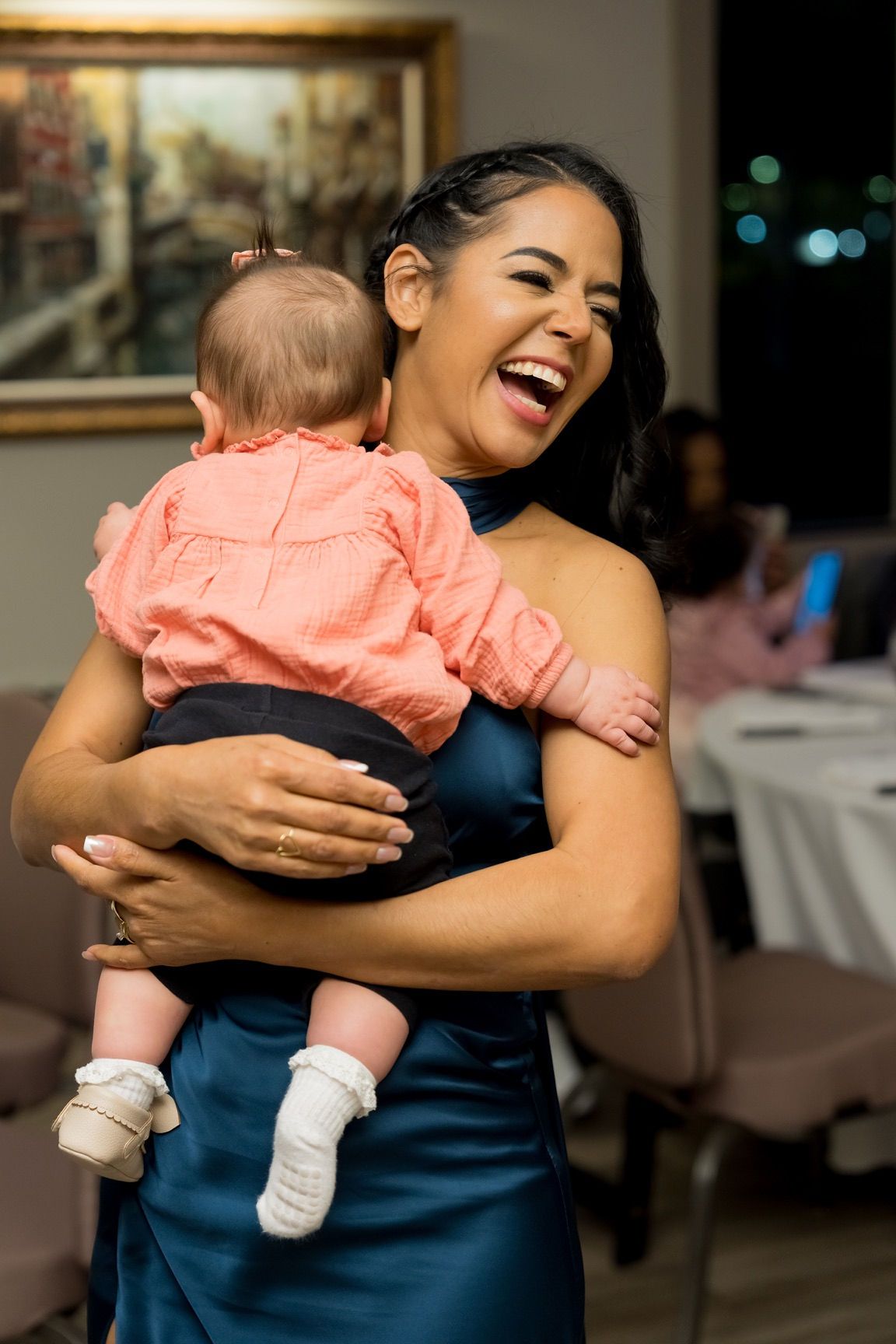 Woman in blue dress laughing while holding a baby wearing pink top and dark shorts.