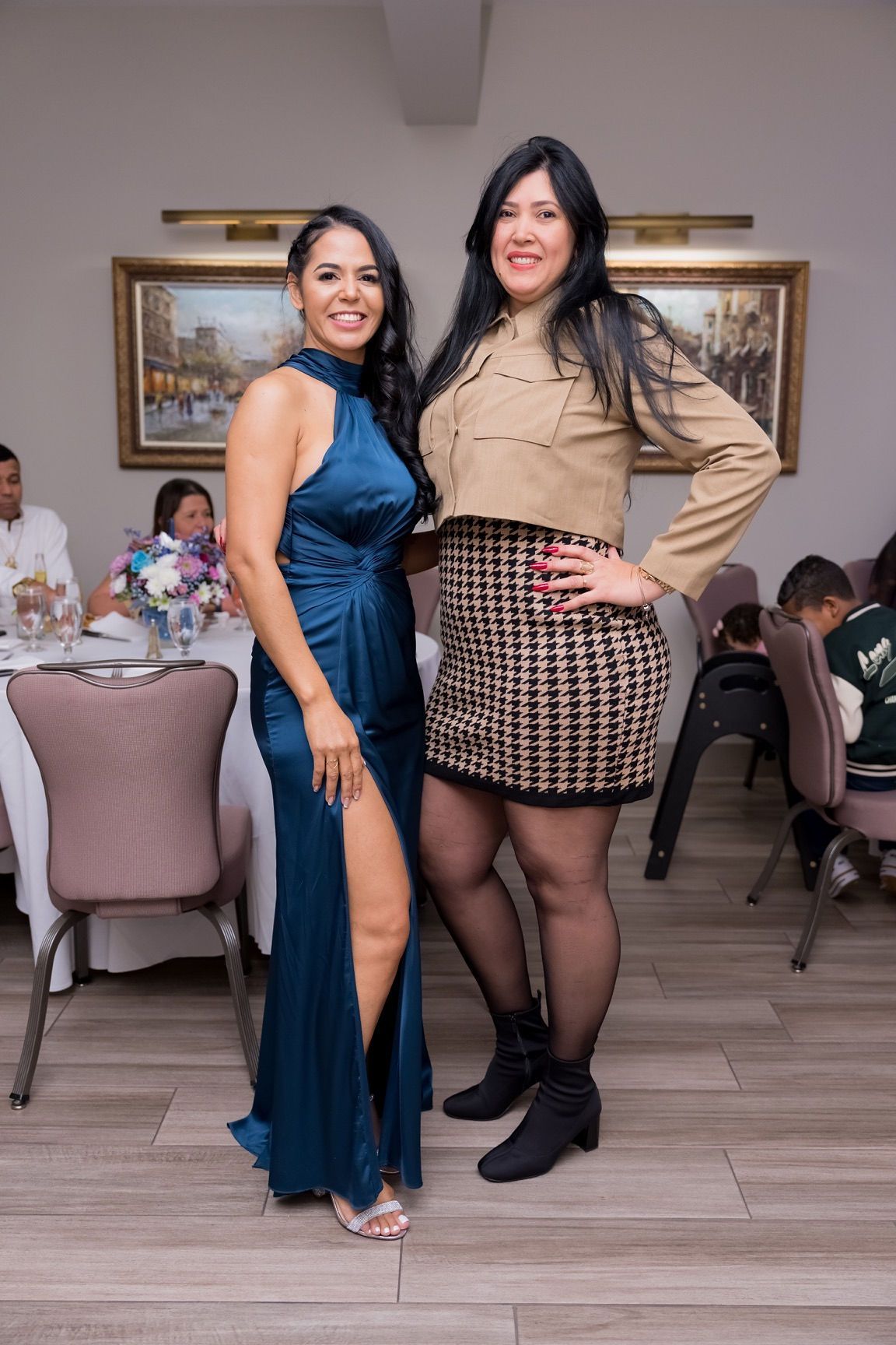 Two women pose at a formal event. One in blue gown, the other in cropped tan jacket and patterned skirt.