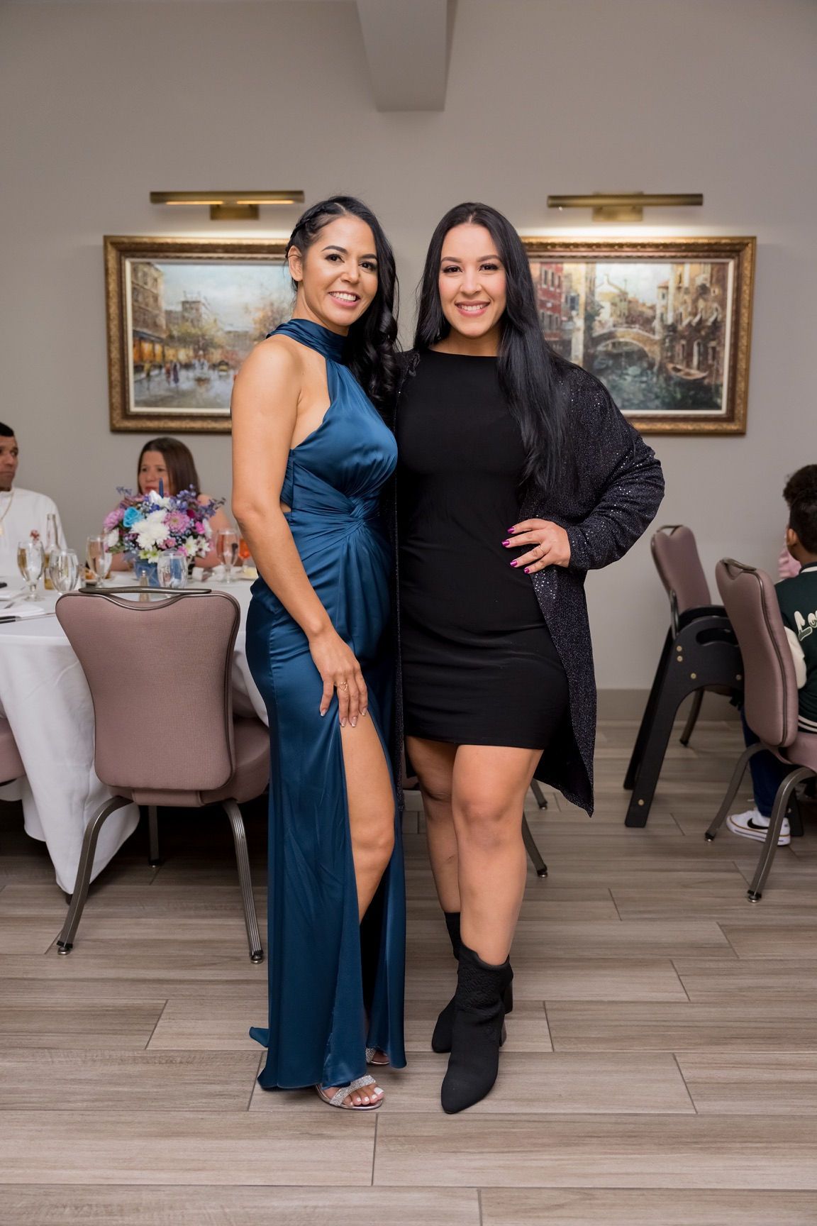 Two women pose together indoors: one in a teal dress with a leg slit, the other in a black dress and sparkly jacket.