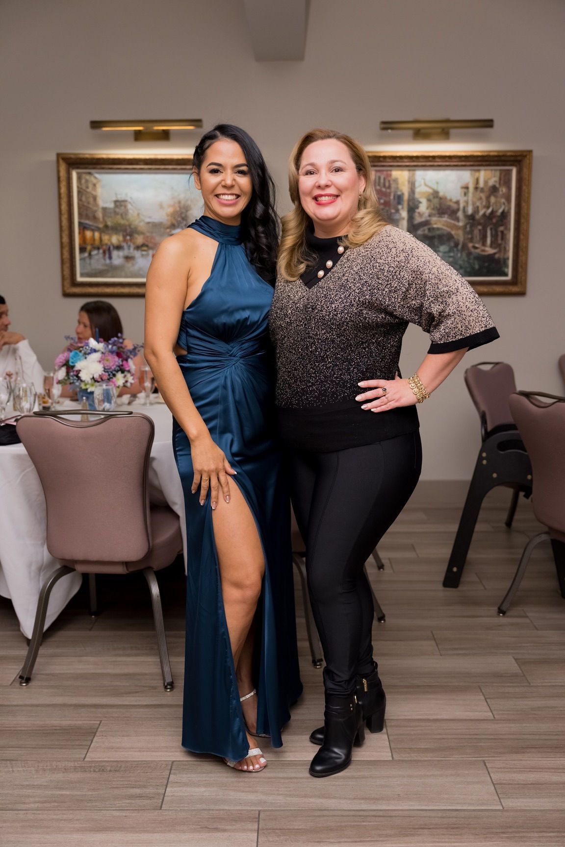 Two women smiling, posing for a photo in a dining hall. One wears a blue gown, the other a patterned top and black pants.