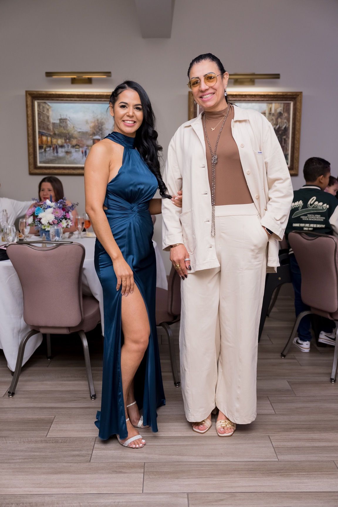 Two women pose at an event. One in blue dress with a slit, the other in a tan outfit.