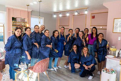 Group of women in blue robes pose for a photo in a beauty salon. Pink walls and decor are visible.