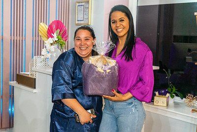 Two women smiling, one in robe holding a gift. The other wears purple top, jeans. Indoors.