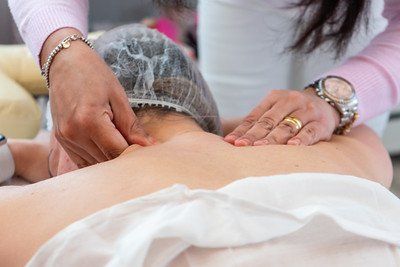 Hands massaging a person's neck and back; the person is lying down, wearing a disposable cap.