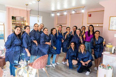 Group of women in blue robes in a pink beauty salon, smiling, some holding drinks.